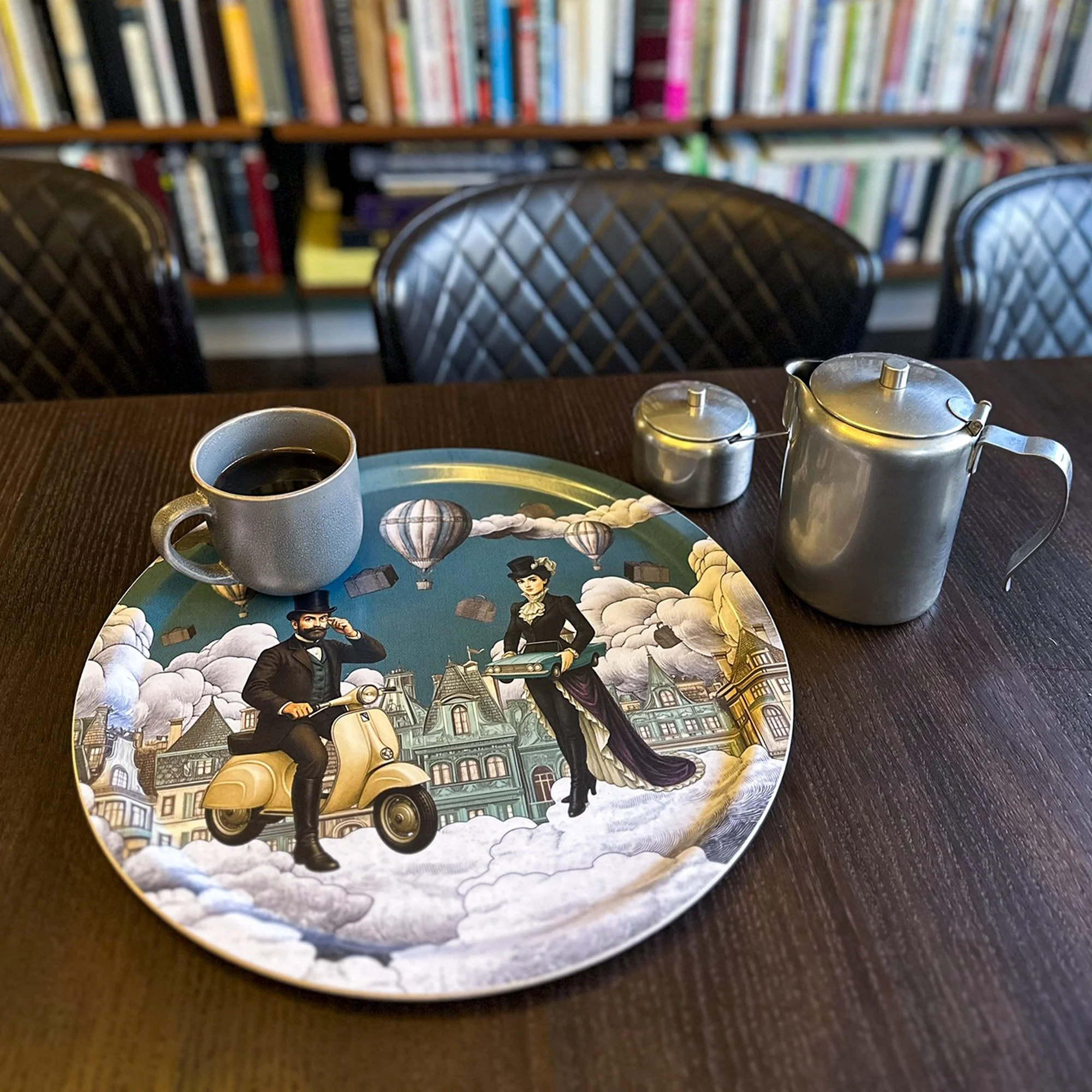 Round birchwood serving tray featuring the 'You Drive Me Crazy' surreal illustration. Depicts a man on a scooter and a woman in clouds with hot air balloons. Designed by Laura Molina for Bontaria.