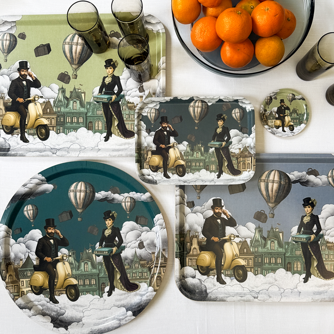 Decorative plates, a glass bowl of oranges, and drinking glasses with a vintage illustration of a man and woman in period clothing riding a scooter among clouds and hot air balloons.