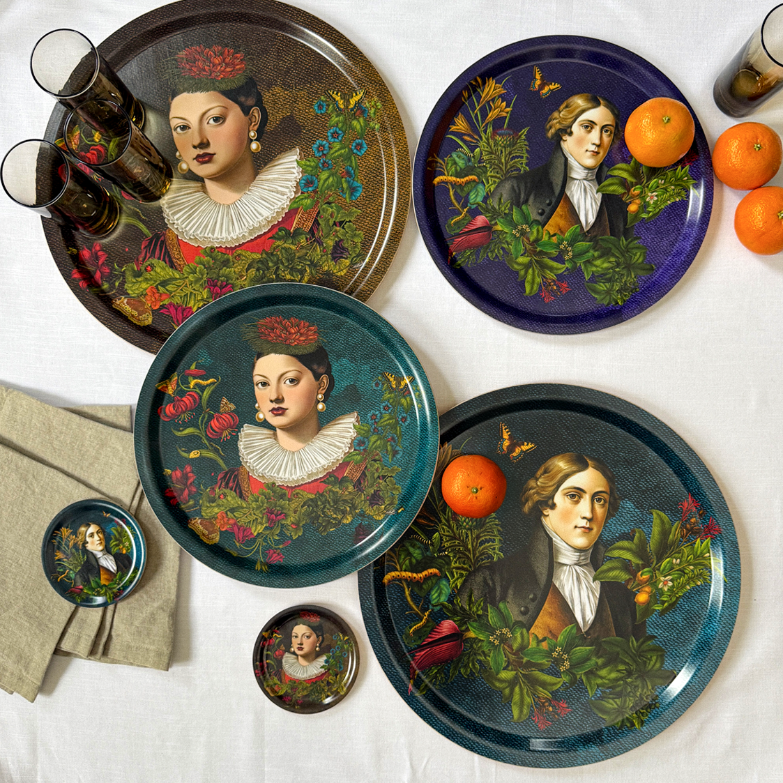 Decorative plates with portraits of a woman with a floral headpiece and a man with a natural background, placed on a white table with oranges and glasses.