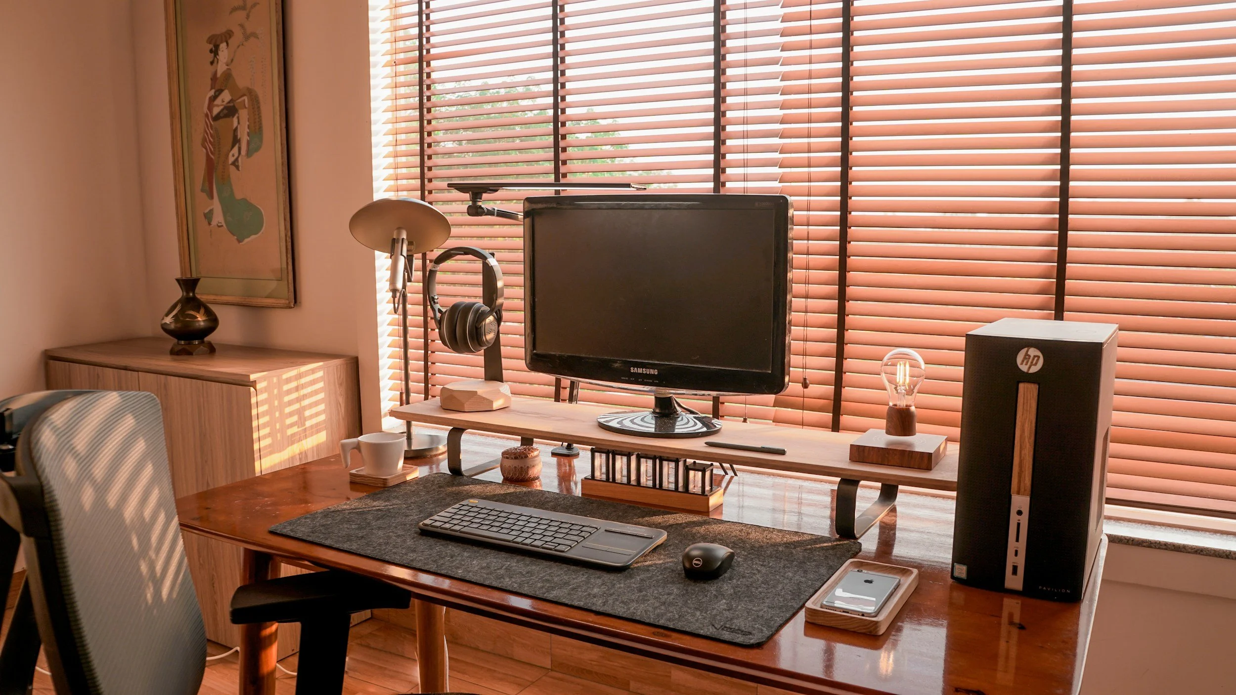 Home office setup with a wooden desk, computer monitor, keyboard, mouse, headphones, lamp, and decorative items, with window blinds in the background.