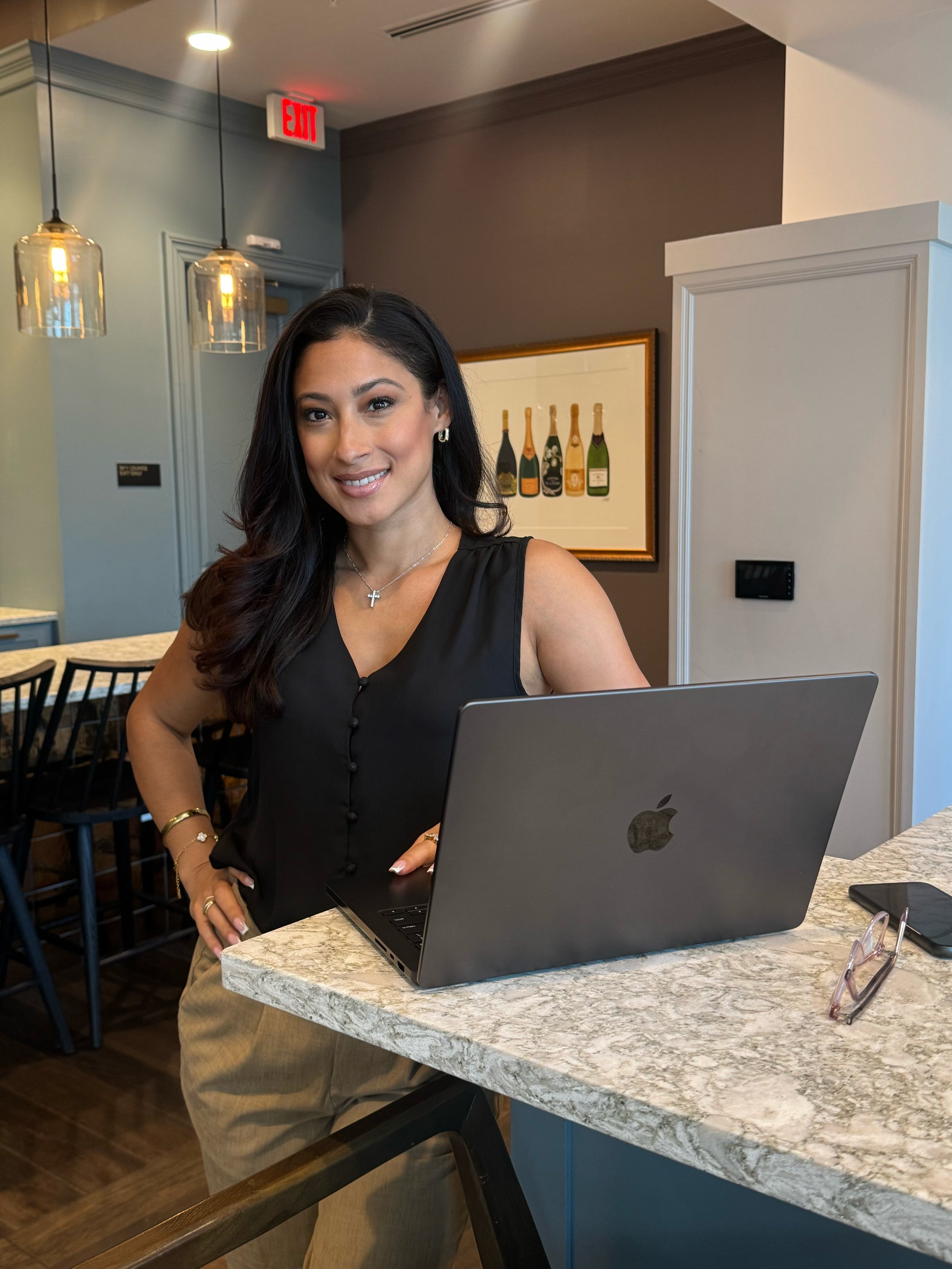 A woman with long dark hair and light skin standing at a kitchen counter with a MacBook laptop, smiling at the camera. She is wearing a black sleeveless top, beige pants, and jewelry. In the background, there are hanging pendant lights, a framed picture of champagne bottles, and a gray wall.
