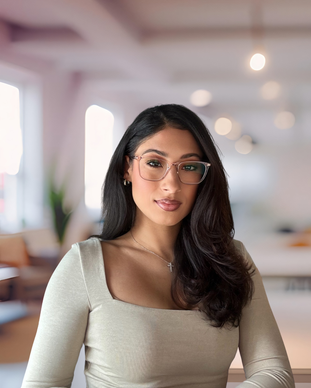 A young woman with long dark hair, wearing glasses and a beige top, standing in a modern, well-lit indoor space with a blurred background.