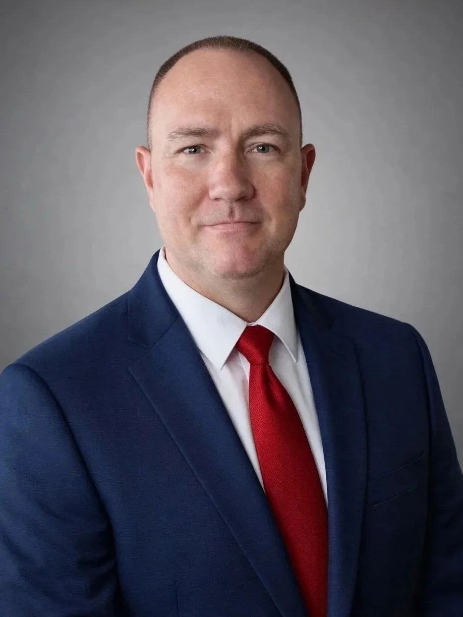 A professional man in a navy suit with a white shirt and red tie, posed against a plain gray background.