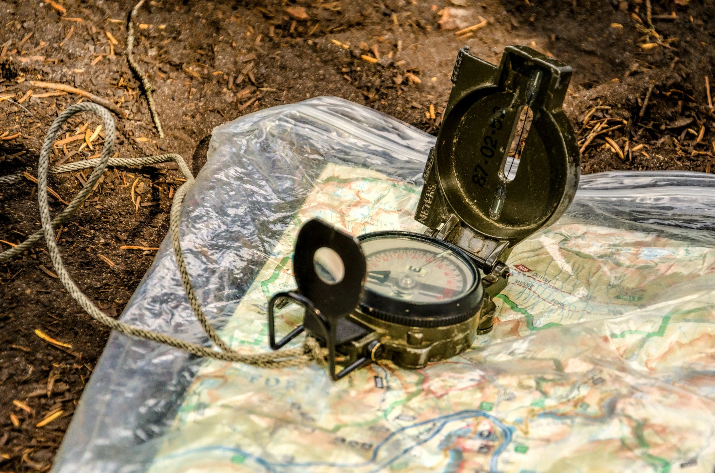 A compass and a map placed on dirt ground, likely in an outdoor or wilderness setting.