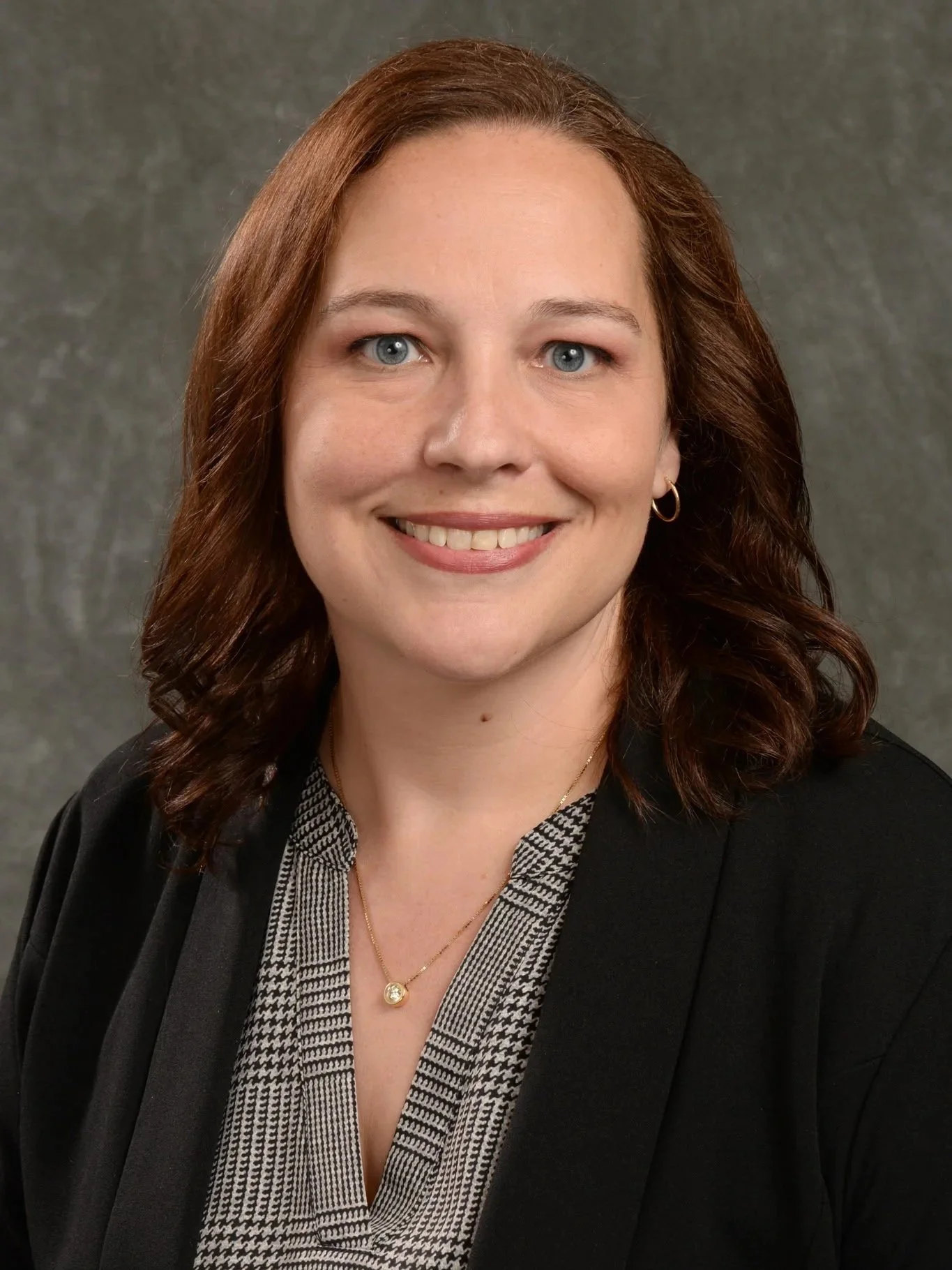 Headshot of a smiling woman with shoulder-length brown hair wearing a black blazer, patterned blouse, and gold jewelry, against a gray background.
