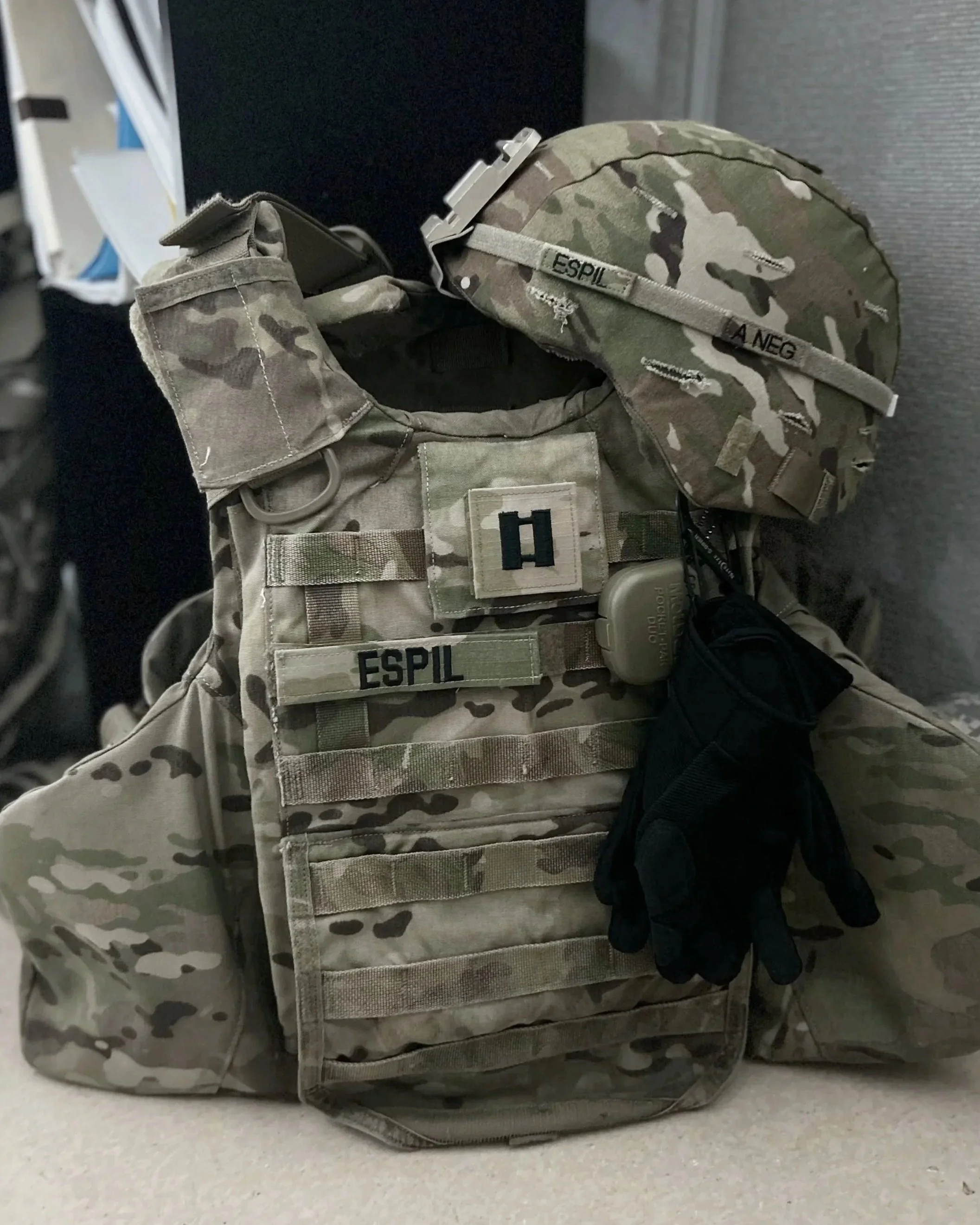Military camouflage tactical vest with name tag 'ESPL,' helmet with camouflage cover, black gloves hanging from the vest, and various pouches and gear attached, set against a neutral background.