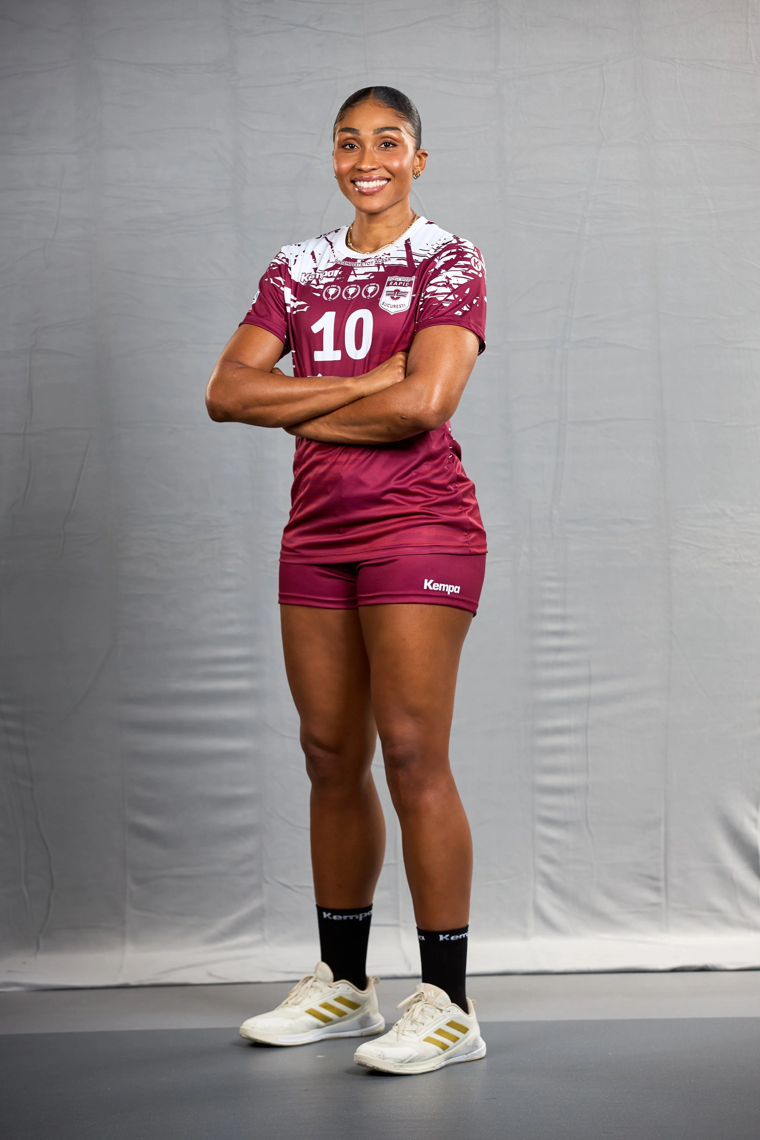 Mulher atleta vestida com uniforme de vôlei, braço cruzado, sorrindo, usando camiseta rosa e shorts, com tênis branco e meias pretas.