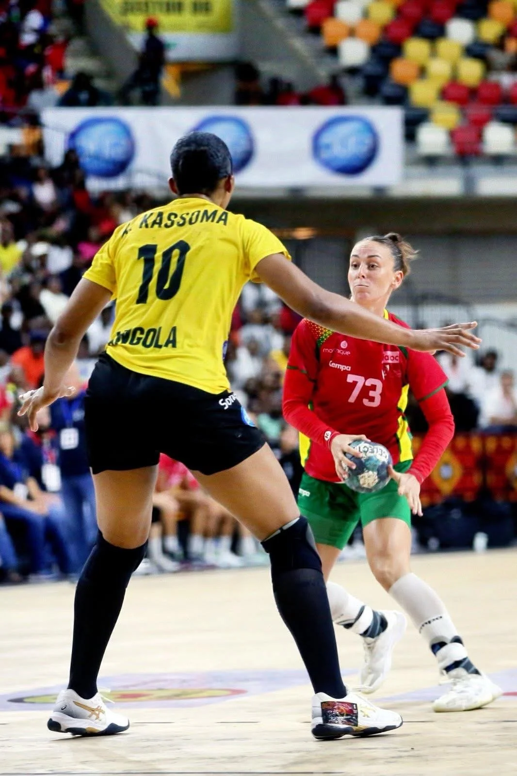 Jogadora de handebol com uniforme amarelo, número 10, enfrentando jogadora adversária de uniforme vermelho, número 73, segurando a bola, em quadra de jogo com público ao fundo.