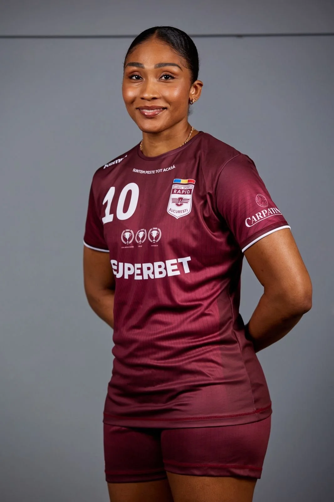 Mulher usando uniforme de time de futebol com camiseta vinho escuro e shorts, sorrindo, com fundo cinza.