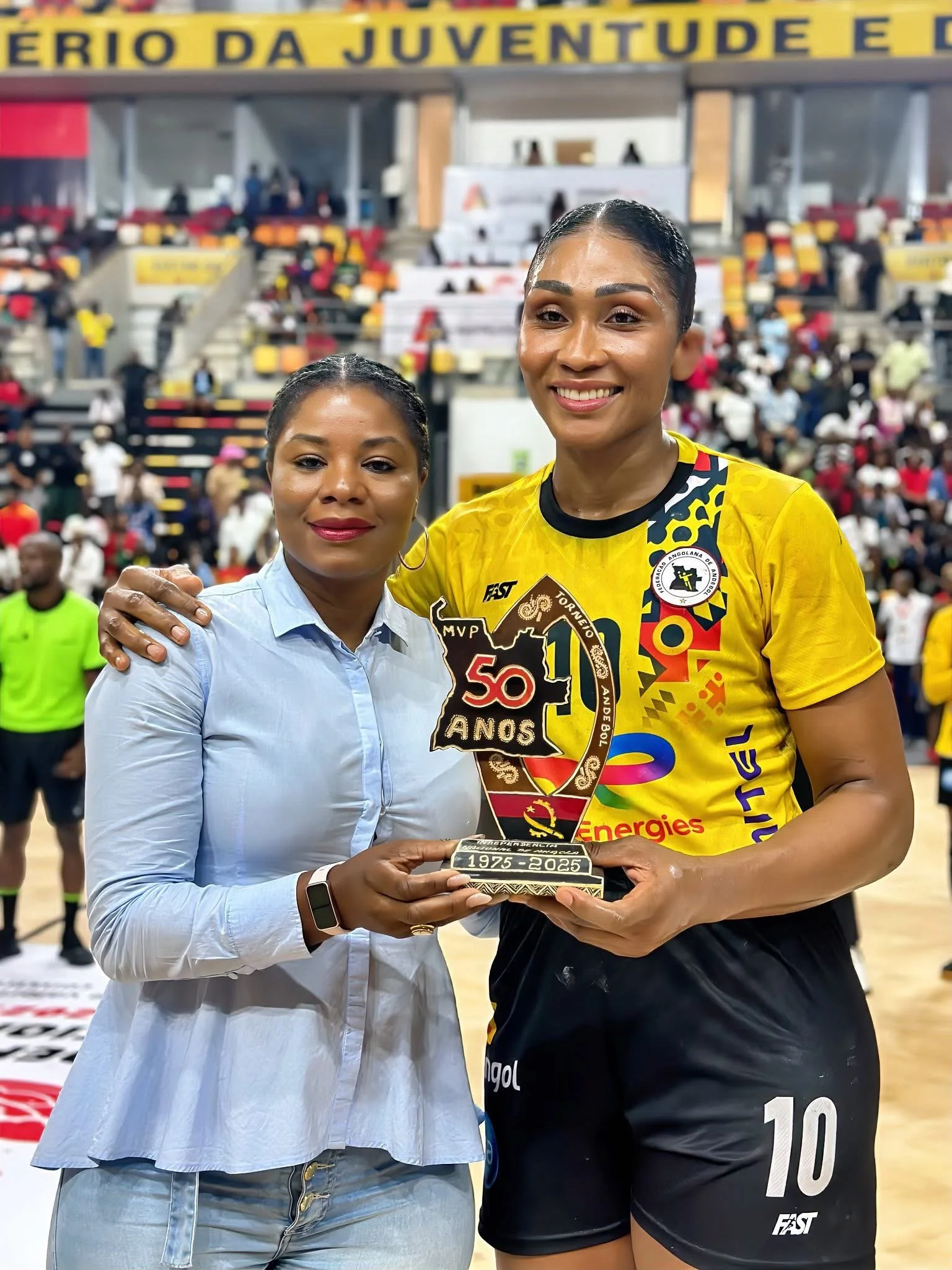 Duas mulheres segurando um troféu dentro de uma quadra de tênis com muitas pessoas ao fundo, comemorando um evento ou vitória.