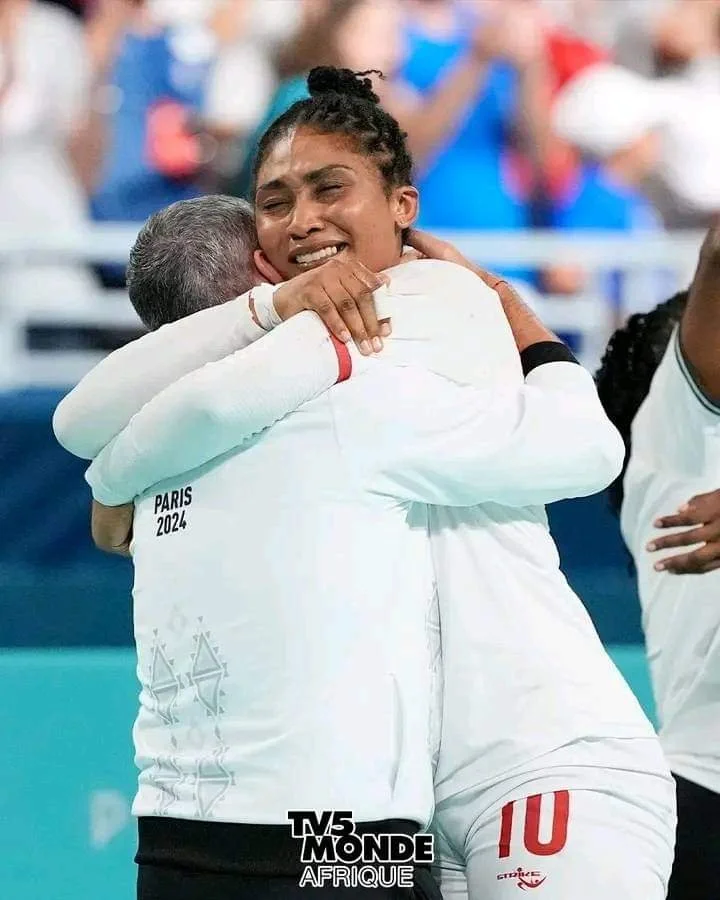 Duas pessoas em um abraço emocionado após uma competição, com fundo de pessoas e uma partida de vôlei. A pessoa da direita está sorrindo e usa uniforme com o número 10.