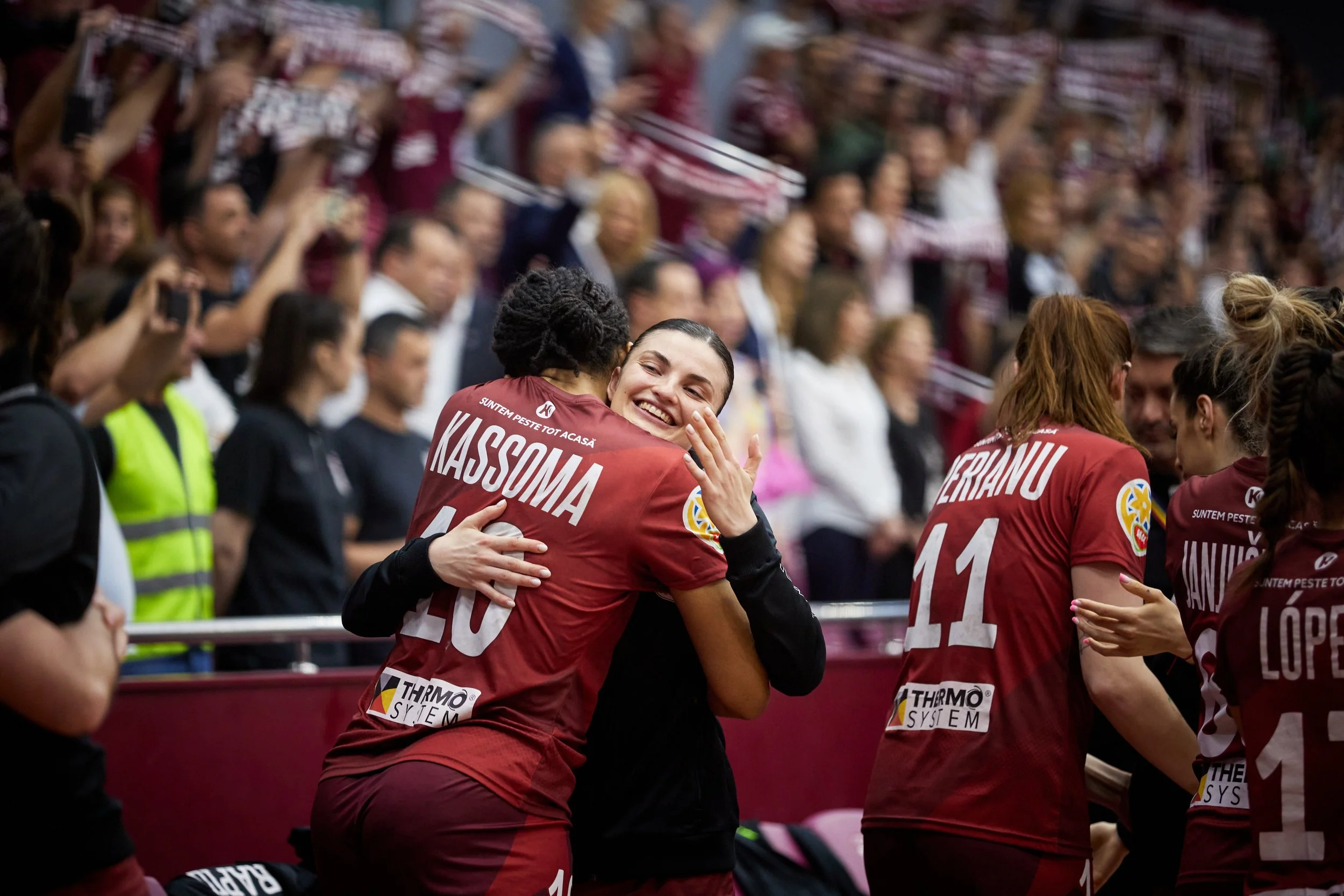 Jogadoras de vôlei comemorando após uma partida, com uma delas abraçando uma pessoa, em uma quadra com torcida ao fundo.
