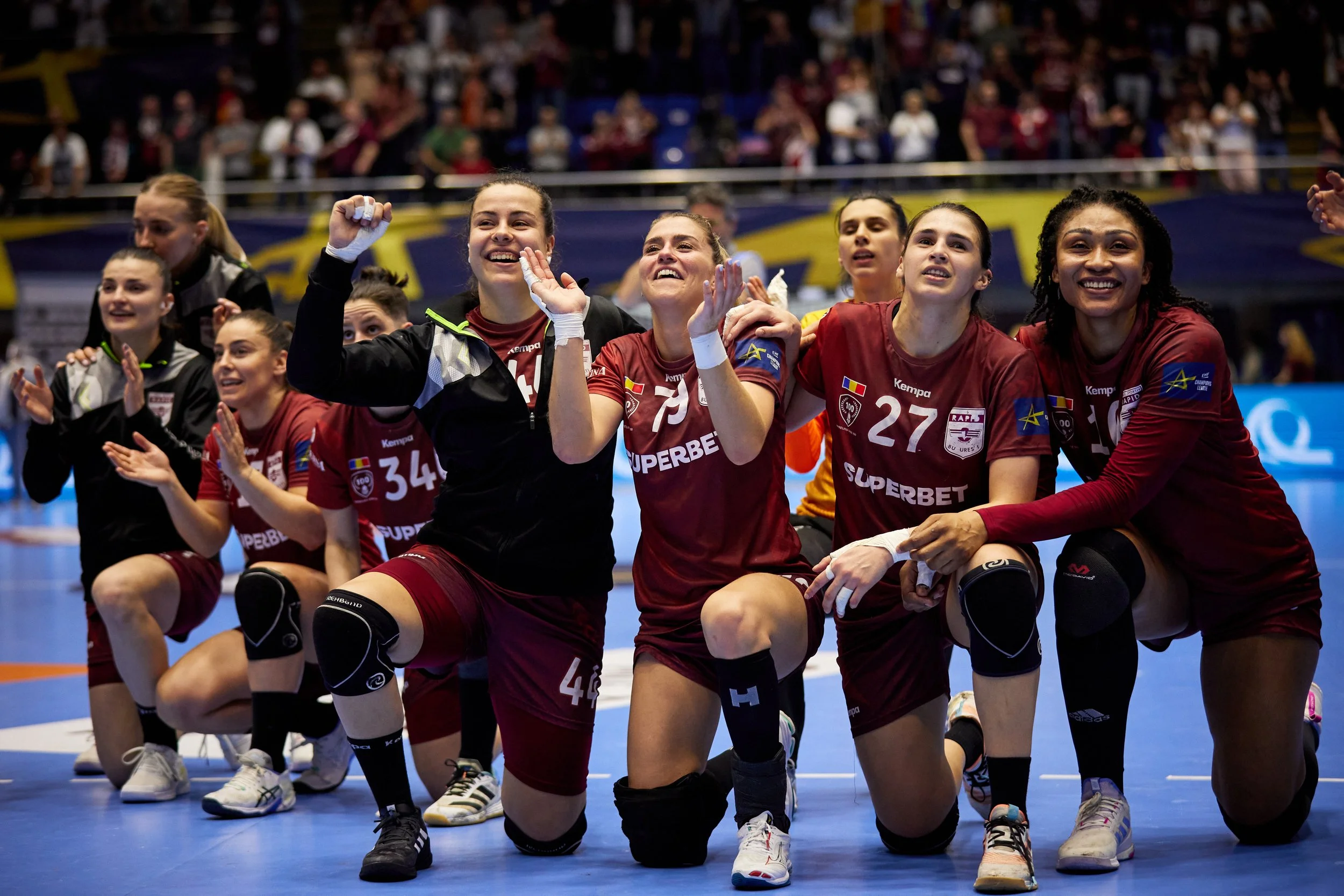 Time de handball feminino comemorando vitória no ginásio, com muitas jogadoras de joelhos e sorrindo, usando uniformes vermelhos e calçados esportivos, ao fundo uma torcida animada.