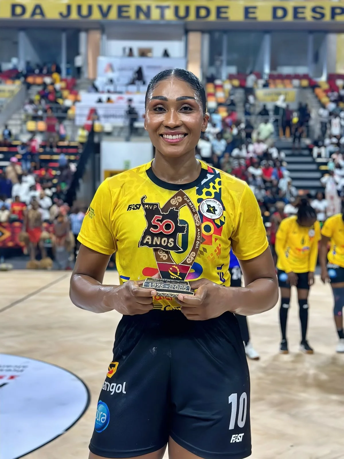 Mulher atleta sorrindo, usando uniforme amarelo e shorts pretos, segurando troféu comemorativo, em quadra de ginástica, com público ao fundo.