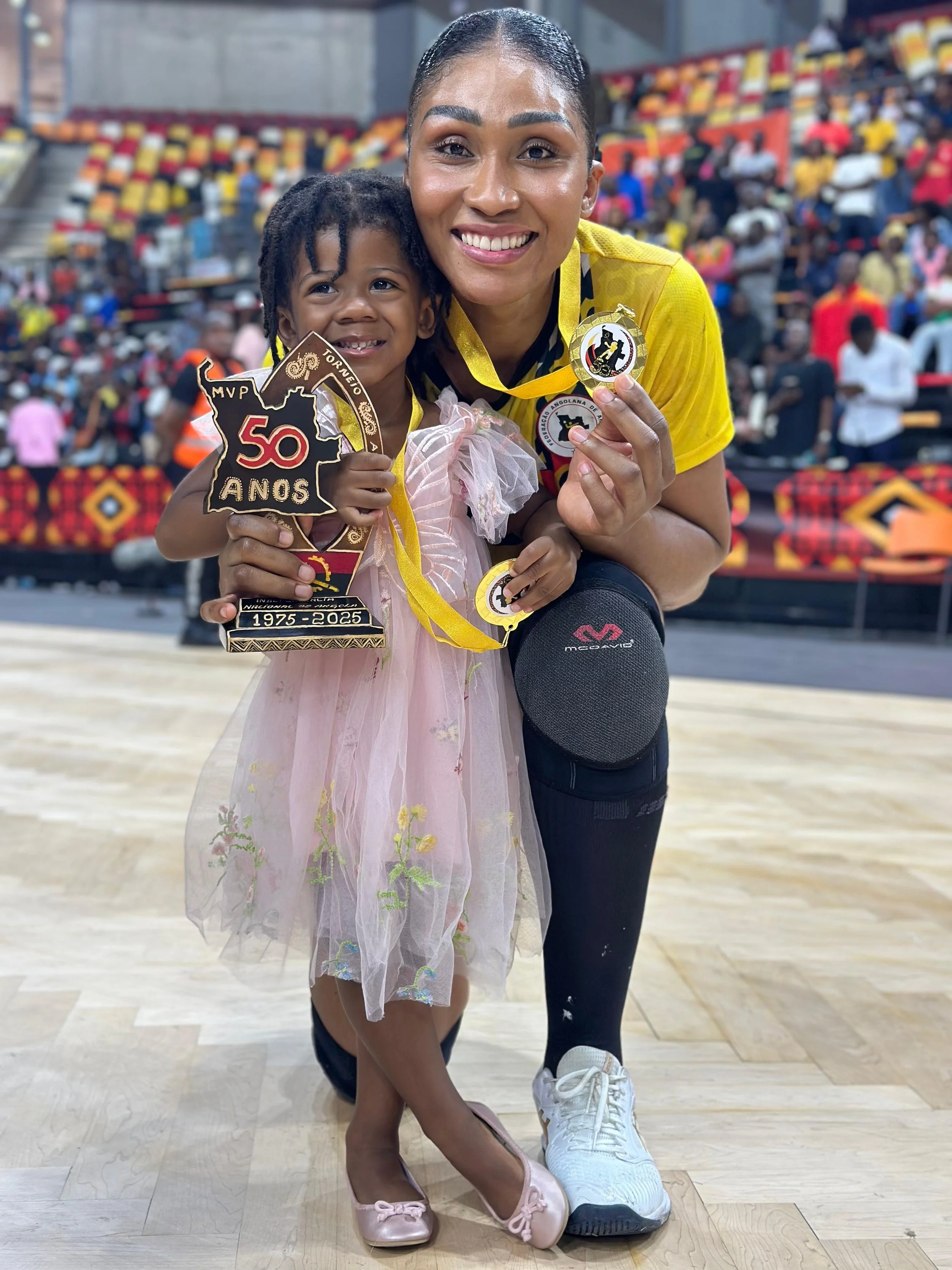Mãe e filha comemorando uma vitória em perfil de uma quadra de esportes, segurando medalhas e um troféu, com público ao fundo.
