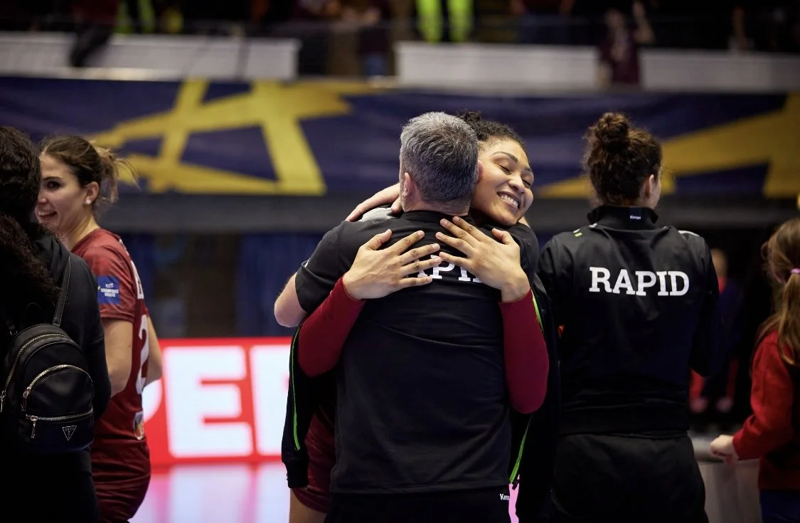 Duas jogadoras de voleibol se abraçando em celebração após um jogo, com outras jogadoras ao redor, em um ambiente esportivo.