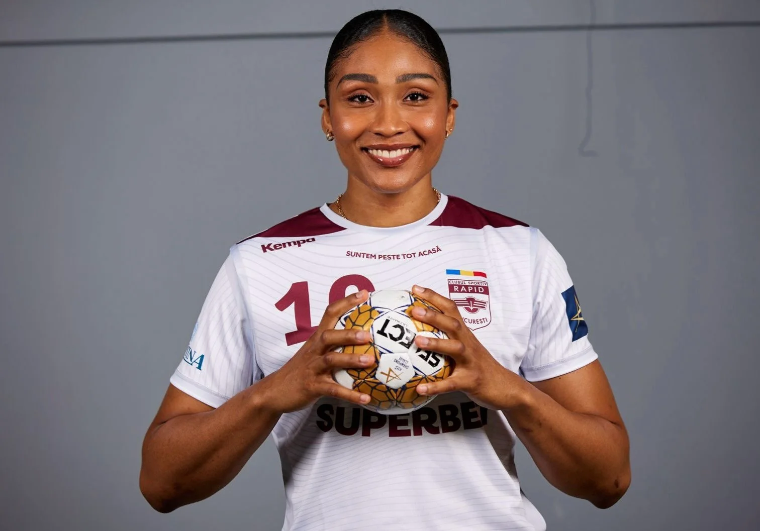 Jogadora de futebol sorridente usando uniforme do time Rapid Bucareste, segurando uma bola de handball com as mãos.