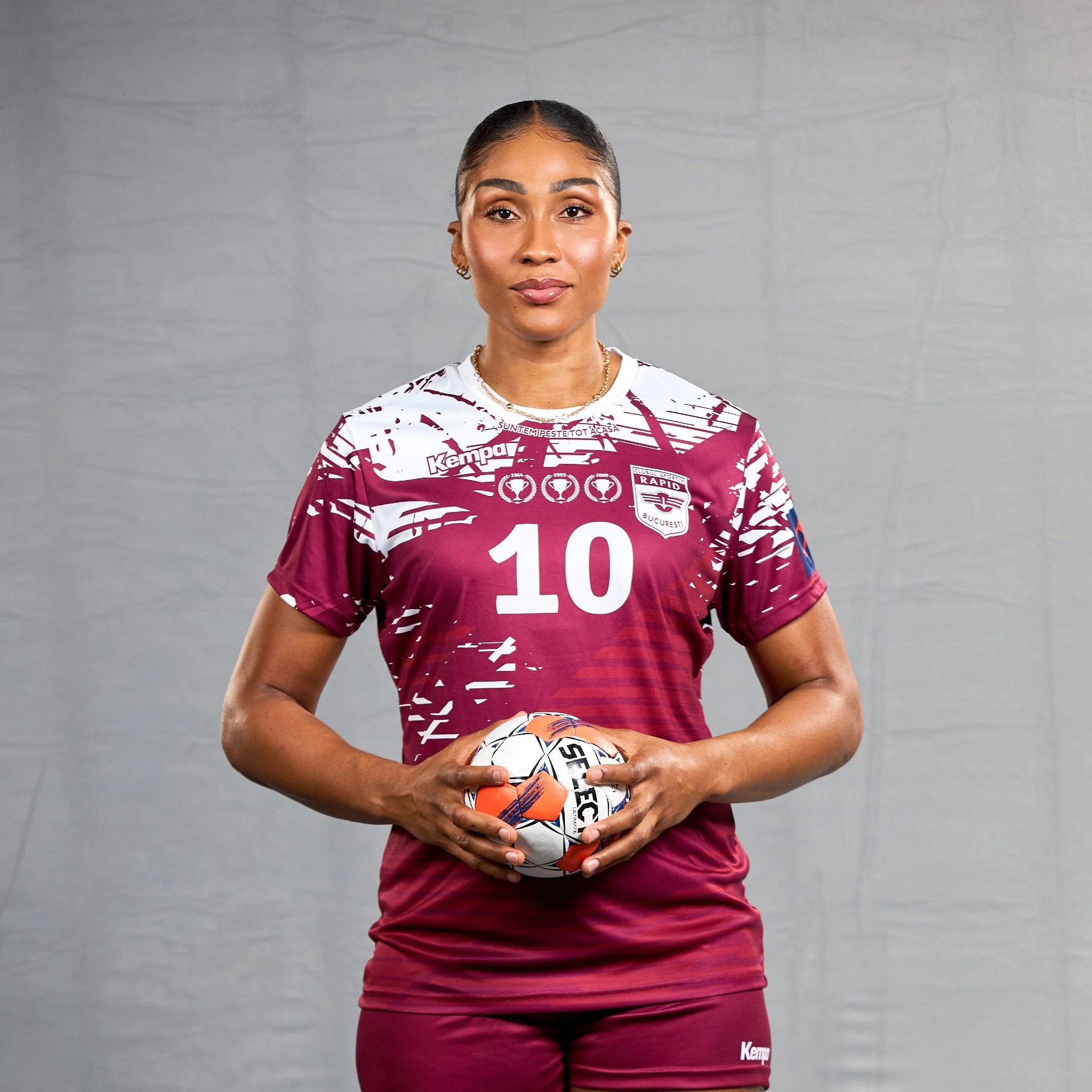 Jogadora de vôlei segurando uma bola, vestindo uniforme de time com o número 10, cor vinho com detalhes brancos.