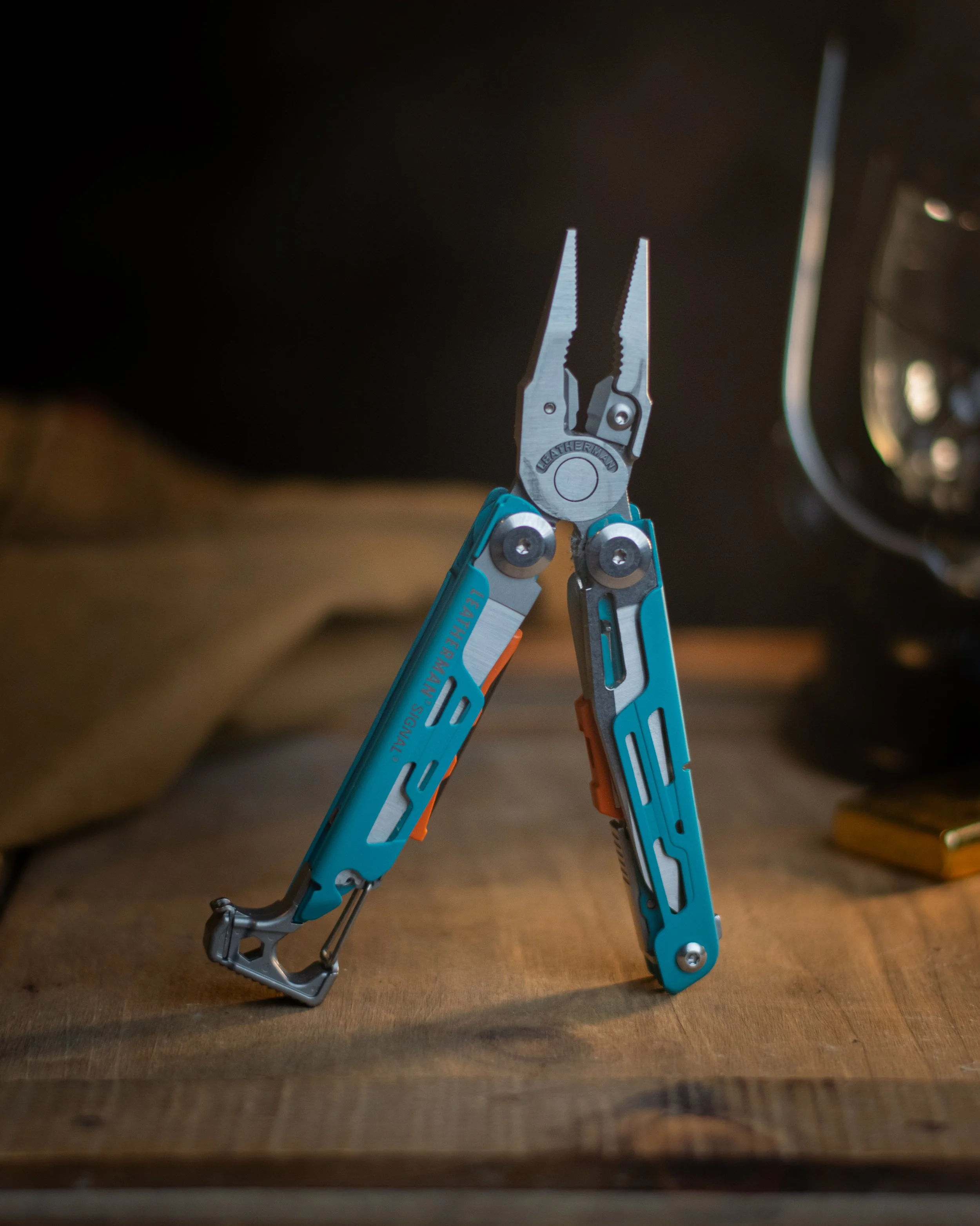 Close-up of a blue multi-tool integrated with pliers, partially opened, resting on a wooden surface. The background is dark and blurred.
