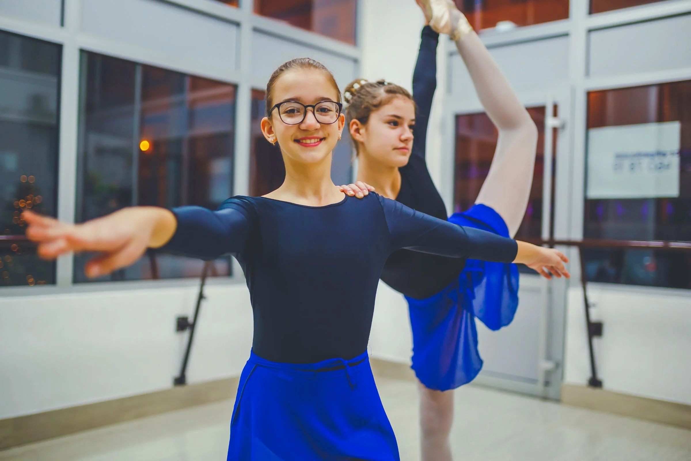 Two students in a dance studio practicing ballet poses and somatic movement near large windows overlooking a city at night.