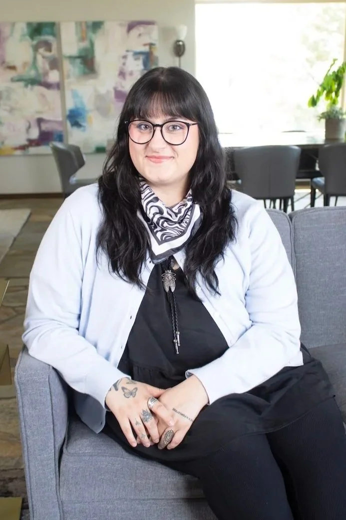 Smiling woman with long dark hair and glasses wearing a light cardigan and patterned neck scarf, seated on a gray couch indoors.