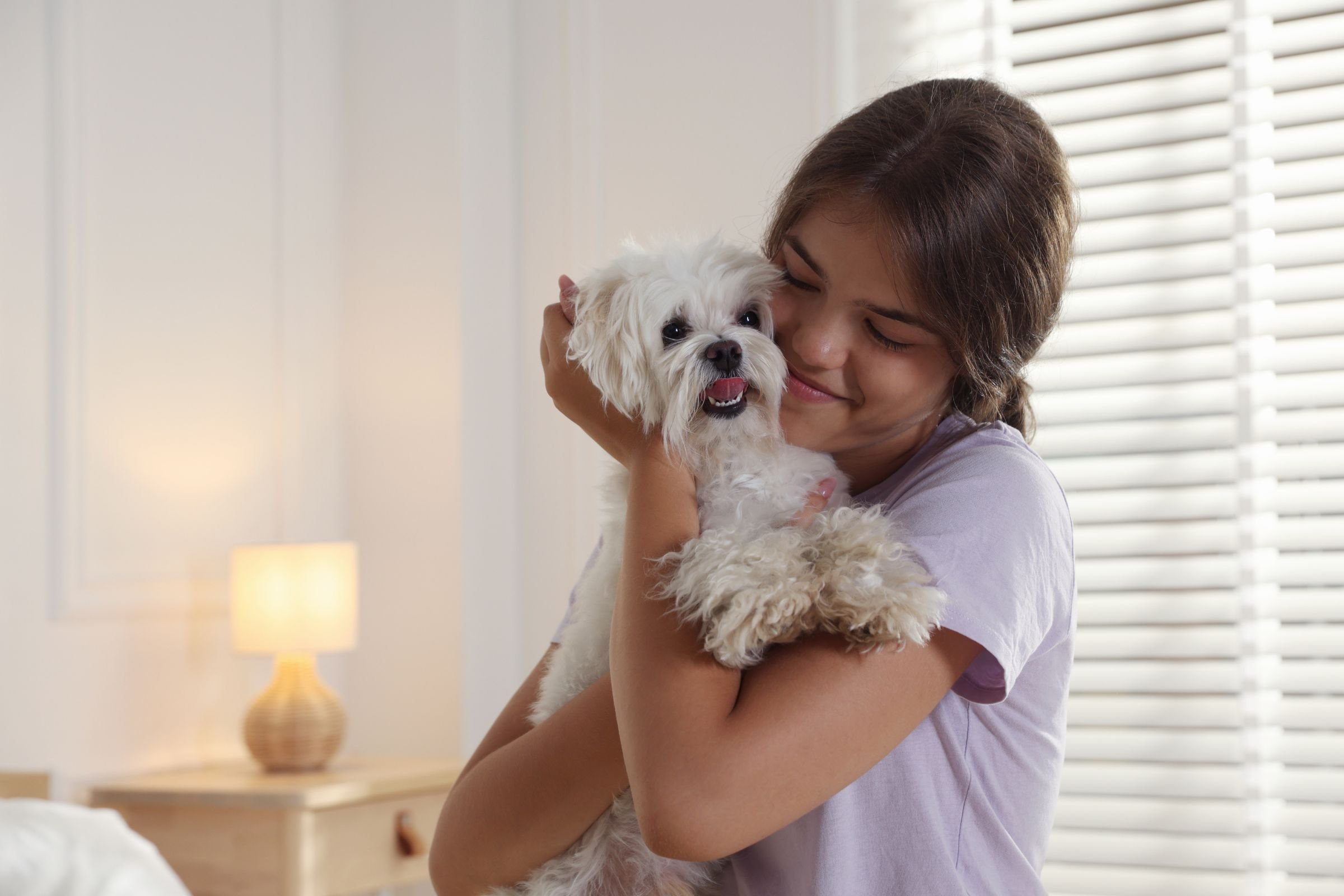 erson hugging a small white dog in a bright room near a window.