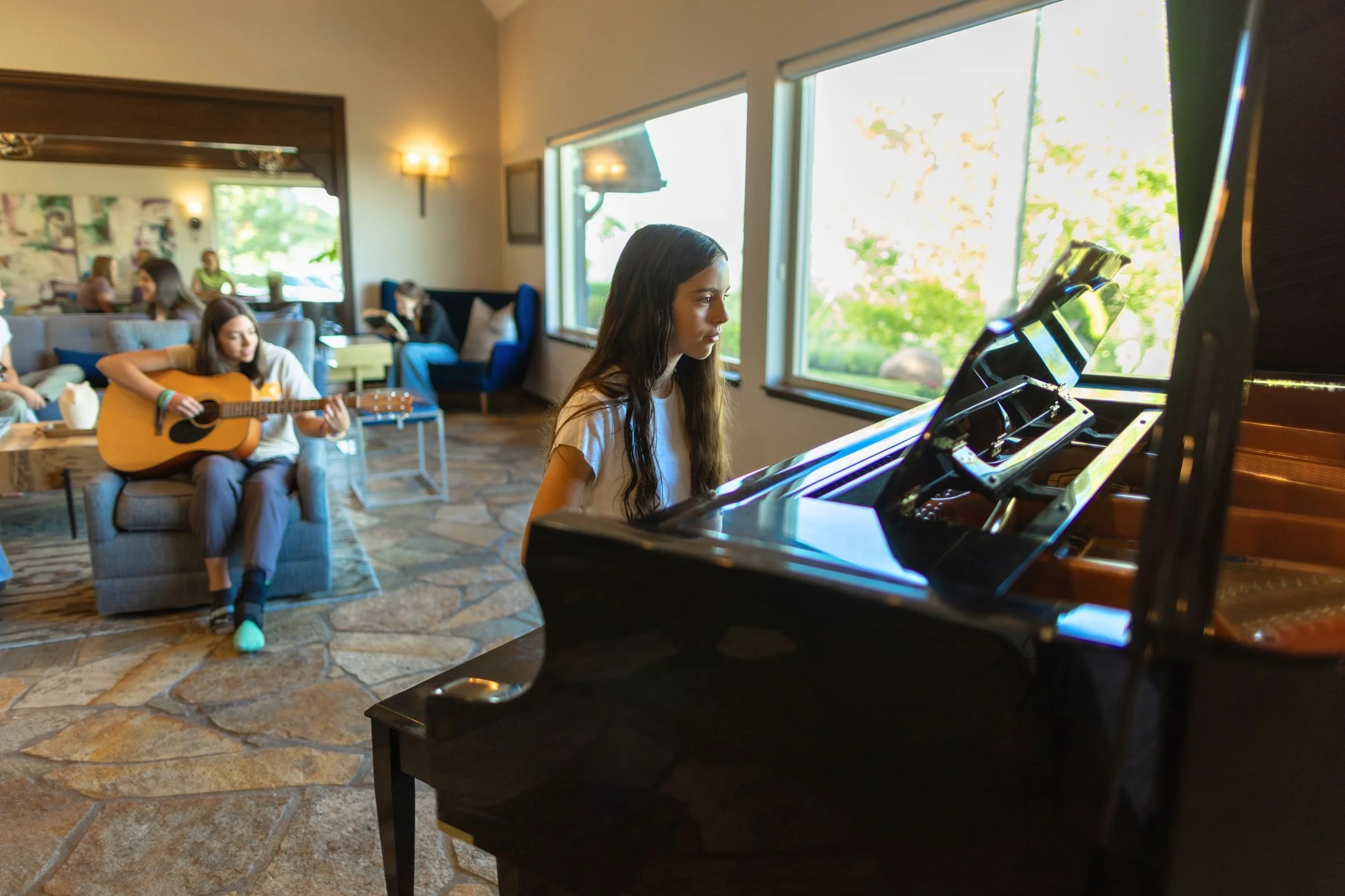 Student plays a grand piano while another plays acoustic guitar in a sunny lounge, with peers nearby.