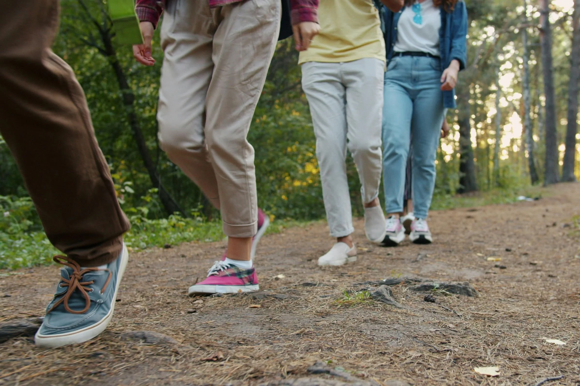 Four people walk on a wooded trail, wearing casual pants and sneakers. The scene conveys a relaxed, social atmosphere in a forest setting.