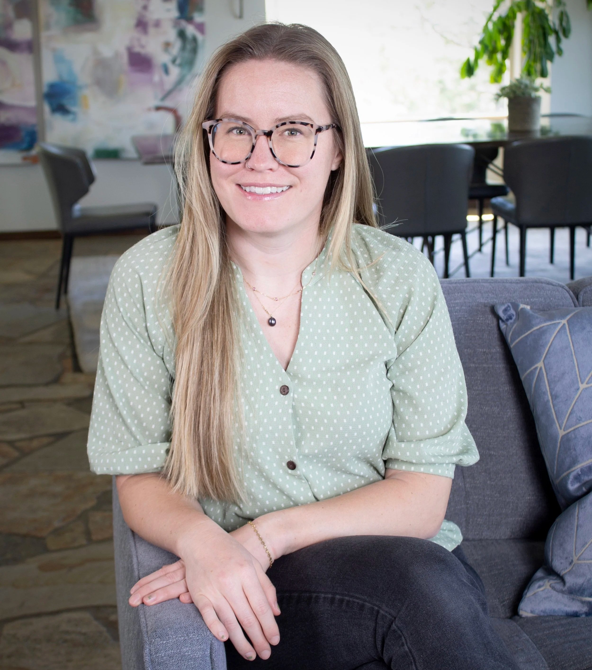 Smiling woman with long blonde hair and glasses, wearing a light green polka-dot blouse and sitting on a gray couch in a bright room.