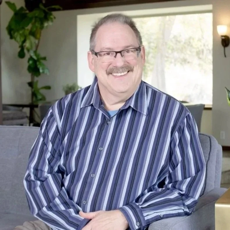 Smiling man with glasses and a mustache wearing a blue striped button-down shirt, seated on a gray couch indoors.