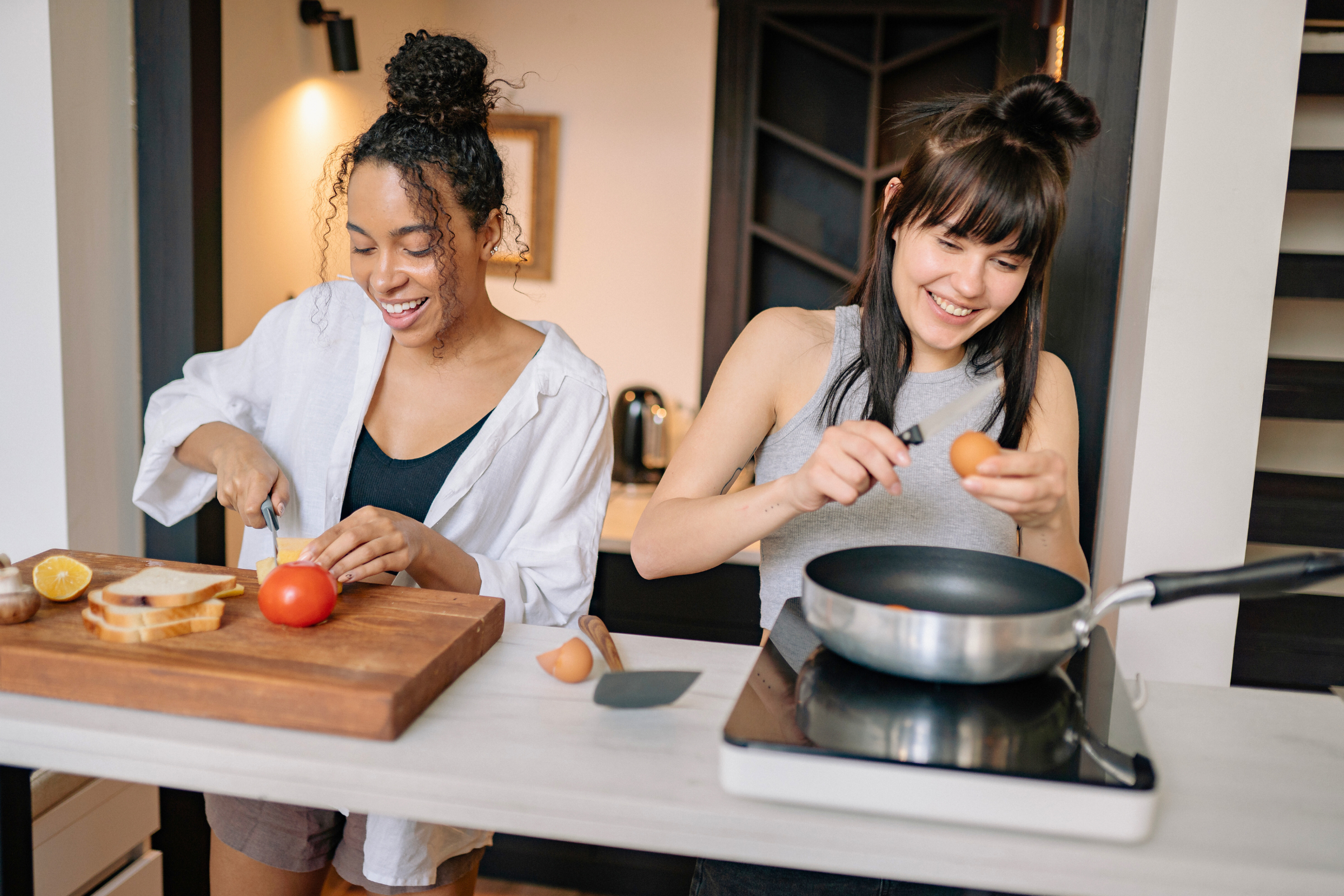 Two young women smile while cooking in a kitchen, one chopping a tomato and one cracking an egg.