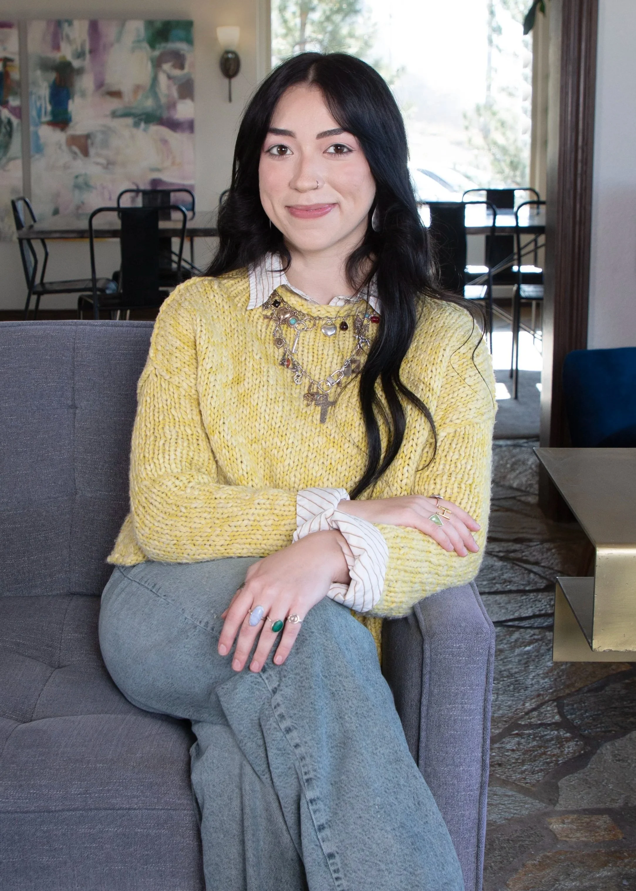 Smiling woman with long dark hair wearing a yellow knit sweater and light jeans, seated on a gray chair indoors.
