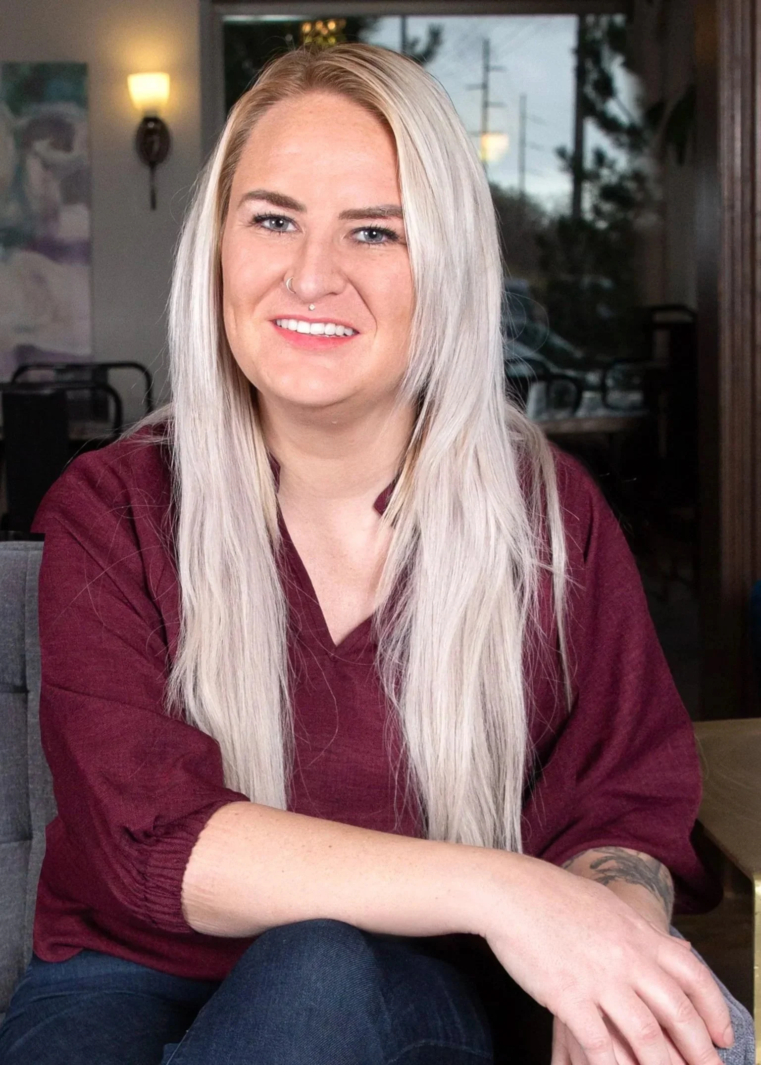 Smiling woman with long platinum-blonde hair, wearing a maroon top and jeans, sitting on a gray couch in a lounge area.