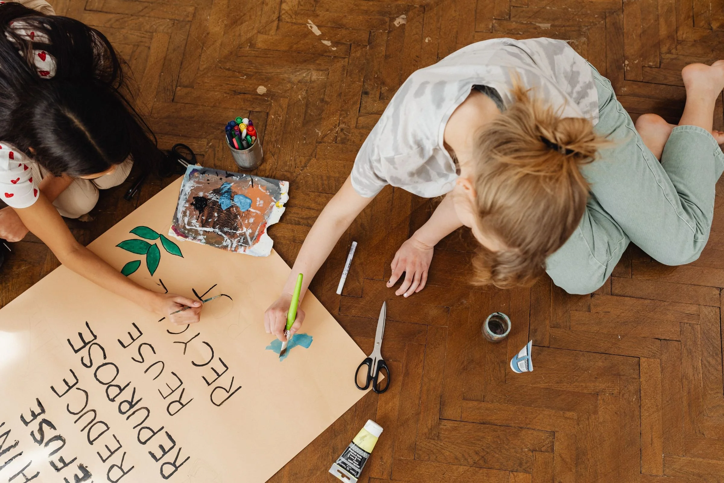 Two young girls sit on a dark wooden floor working together on a craft paper poster painted with environmental slogans and green leaves.