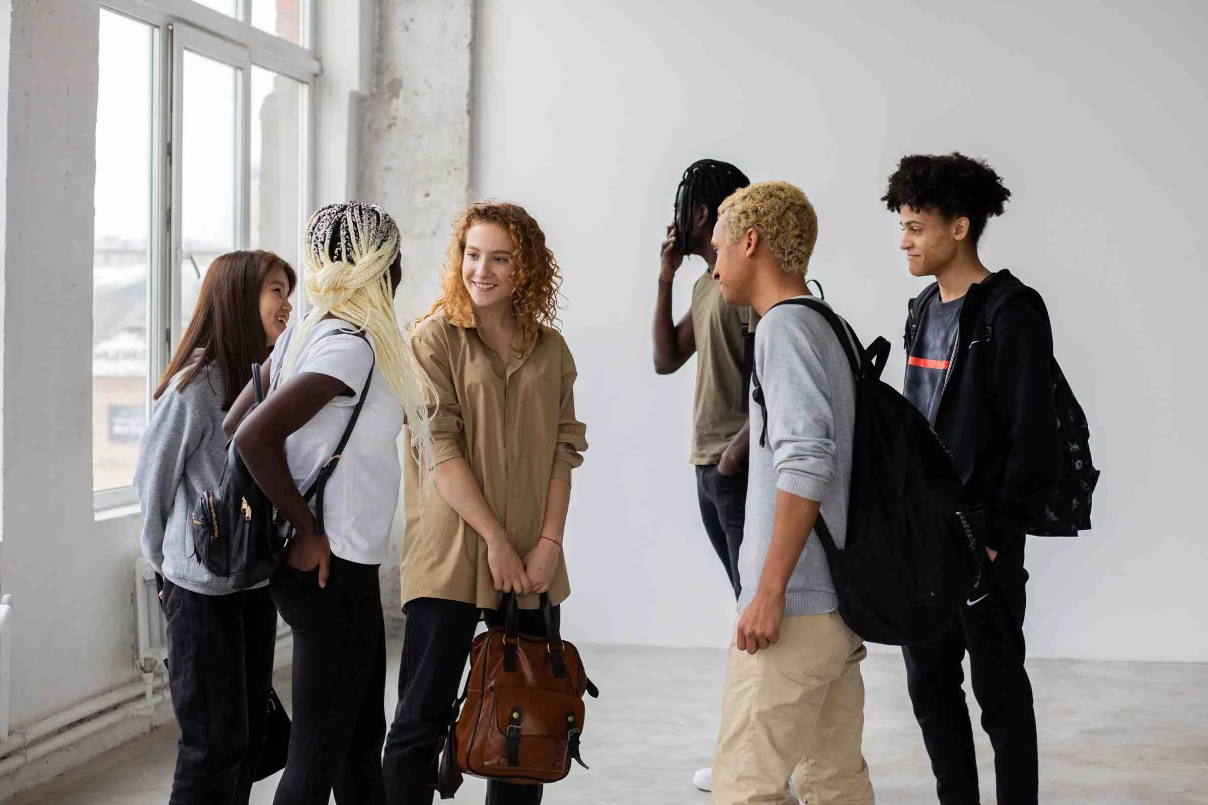 A diverse group of six young adults laughing and chatting in a bright room. They wear casual clothing and have backpacks, suggesting a relaxed, friendly gathering.