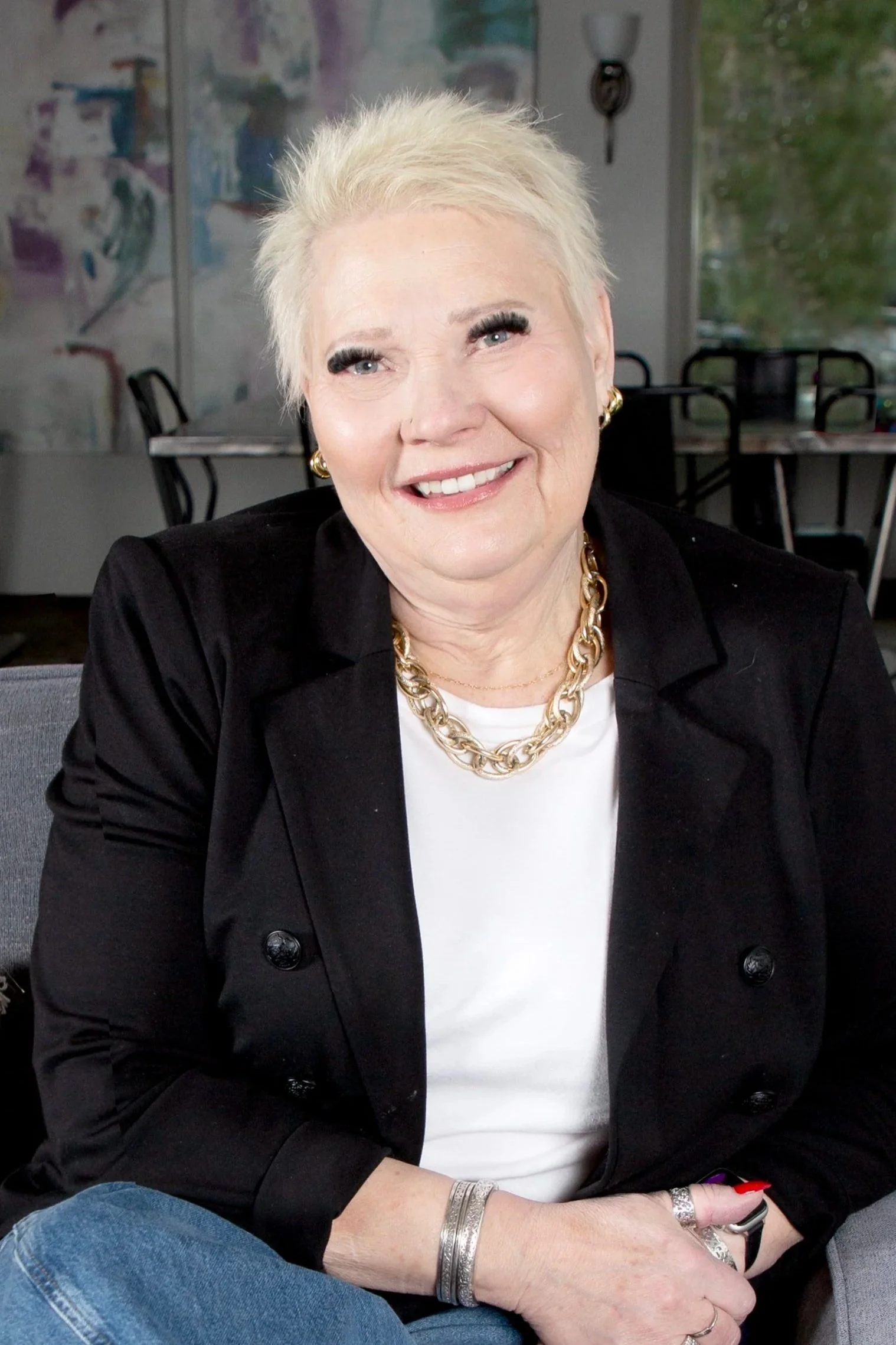 Smiling woman with short blonde hair in a black blazer and jeans, seated on a gray sofa in a bright office.