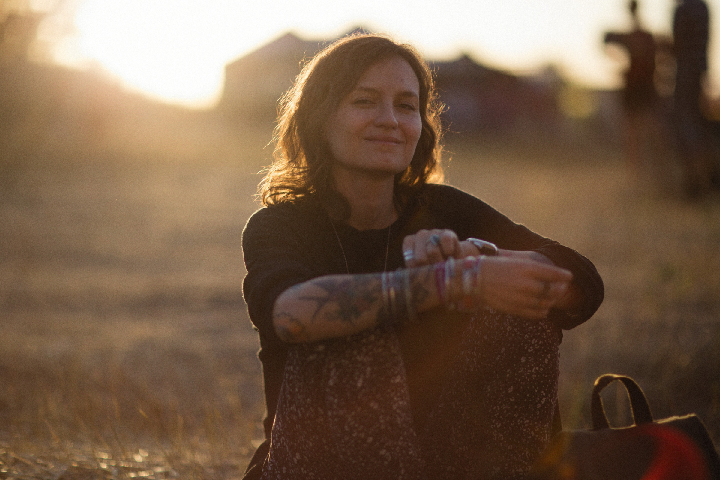 Person sitting in an open field during golden hour, relaxed posture with arms around knees, warm sunlight and a softly blurred background.