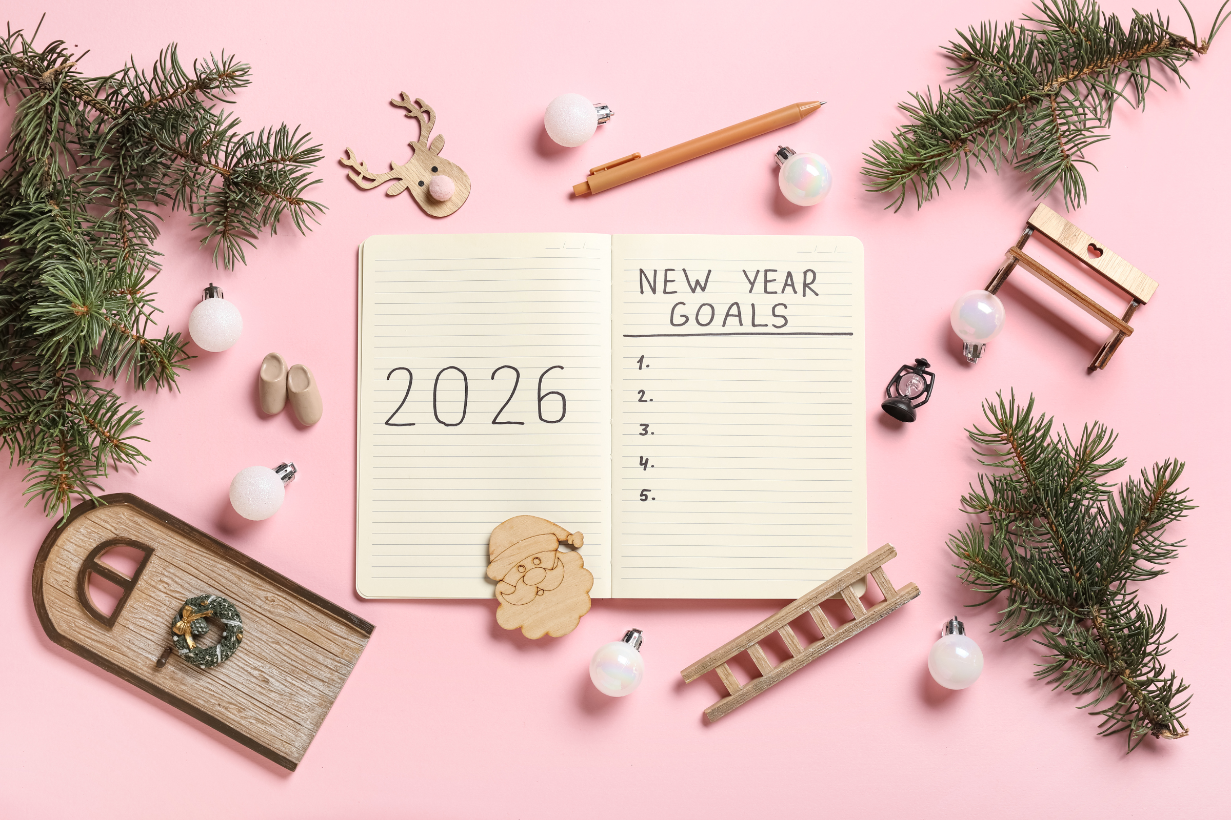 Open notebook reading “2026” and “New Year Goals” on a pink background, surrounded by fir sprigs, ornaments, small wooden decorations, and a pen.