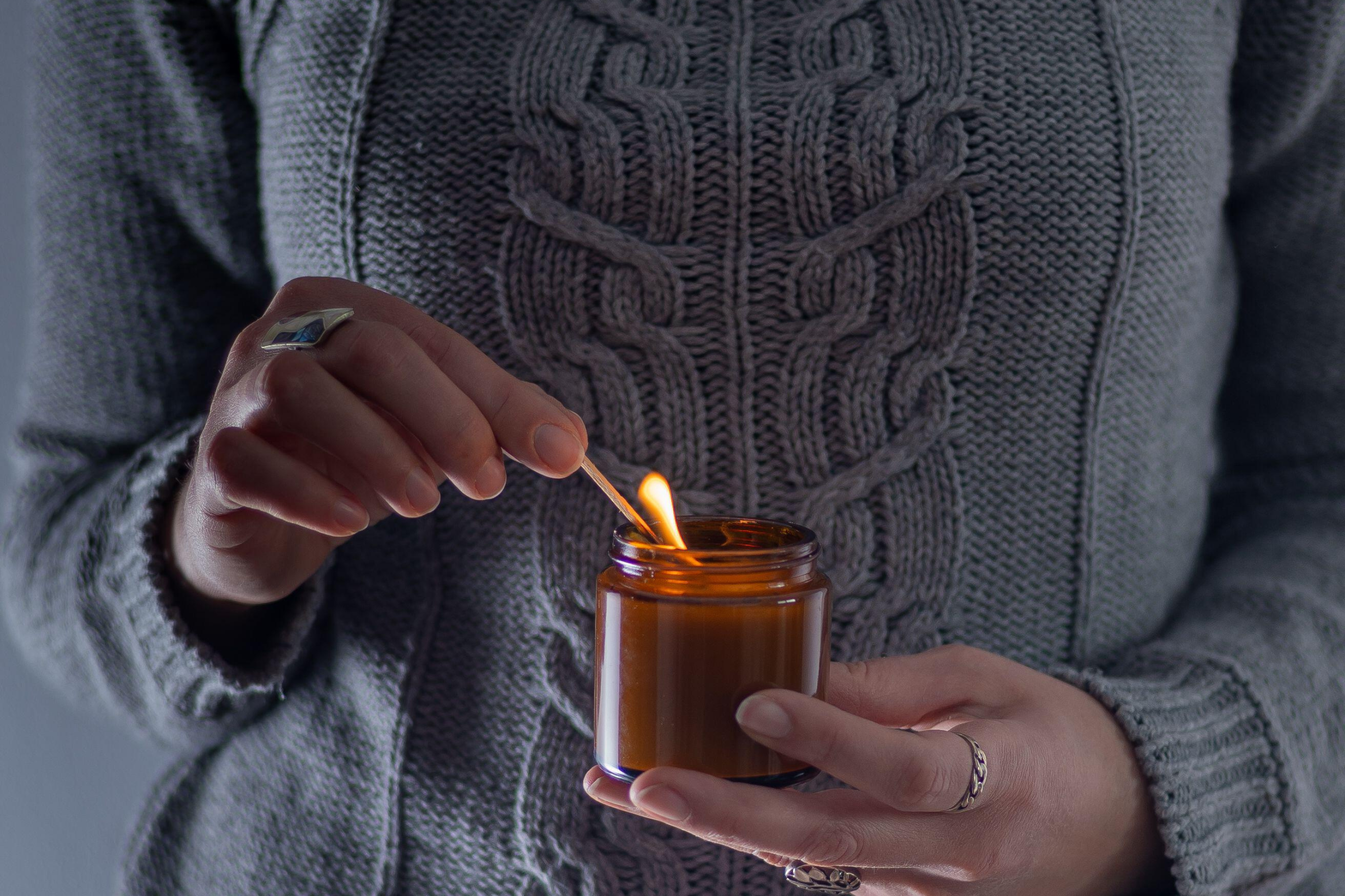 Hands in a gray cable-knit sweater lighting a small amber glass candle with a match.