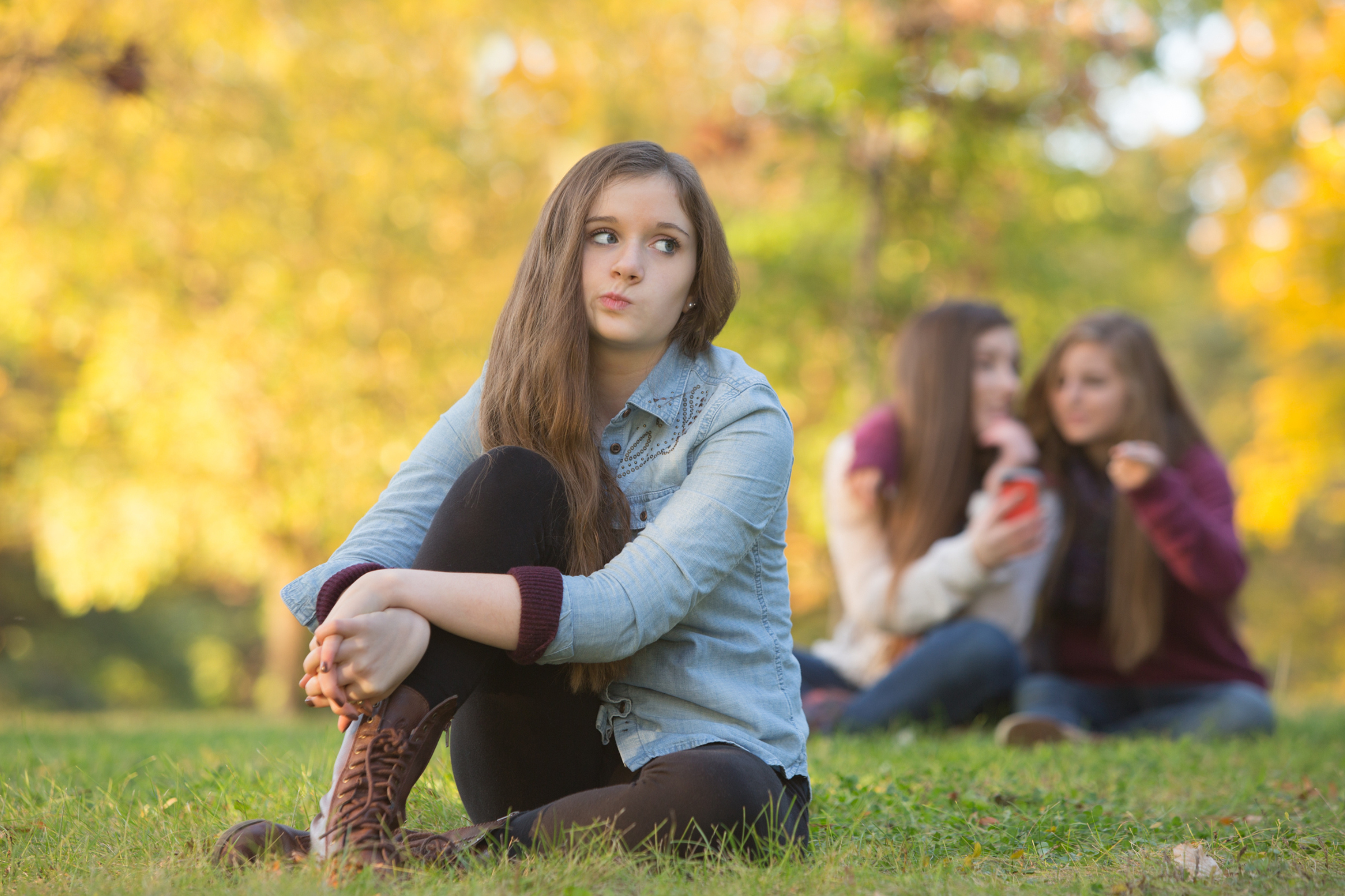 Teen girl sitting on grass outdoors, looking off to the side, with two girls talking in the background.