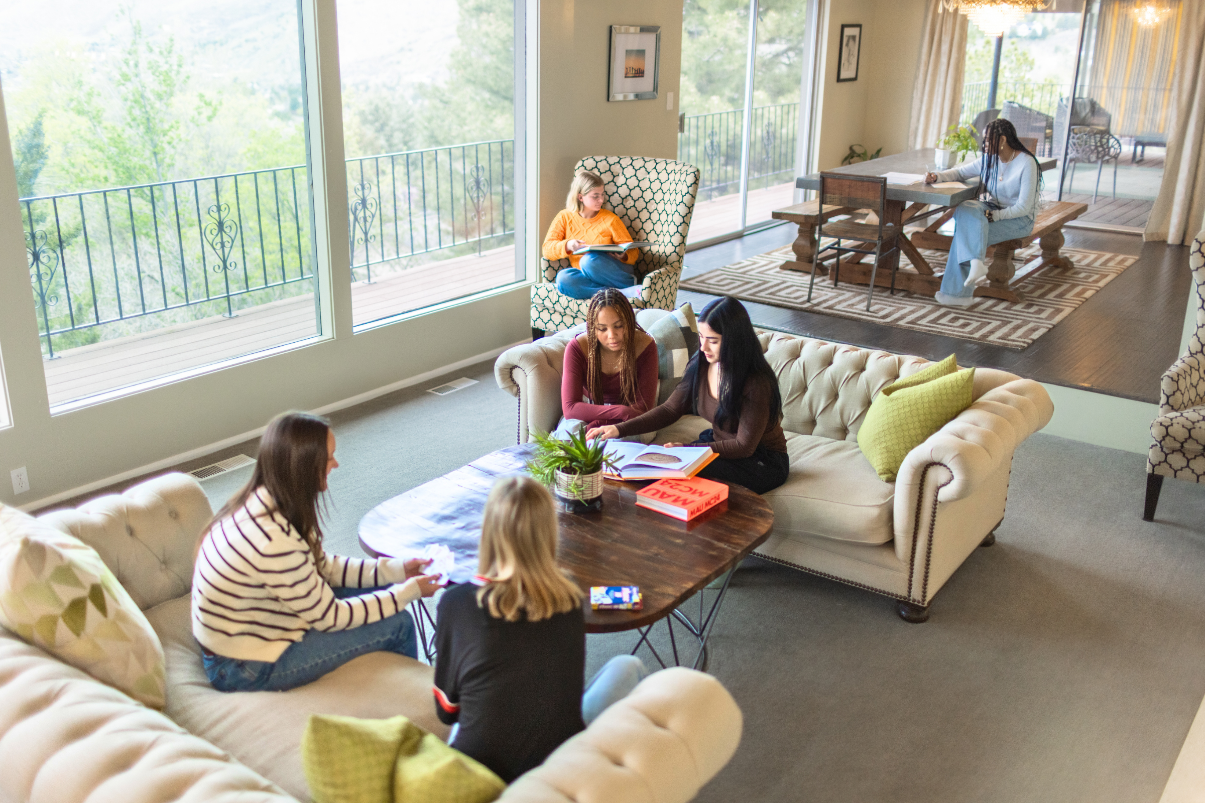 Students read and study in a bright, home-like living room with sofas and a dining table, framed by large windows looking out to a mountain view.