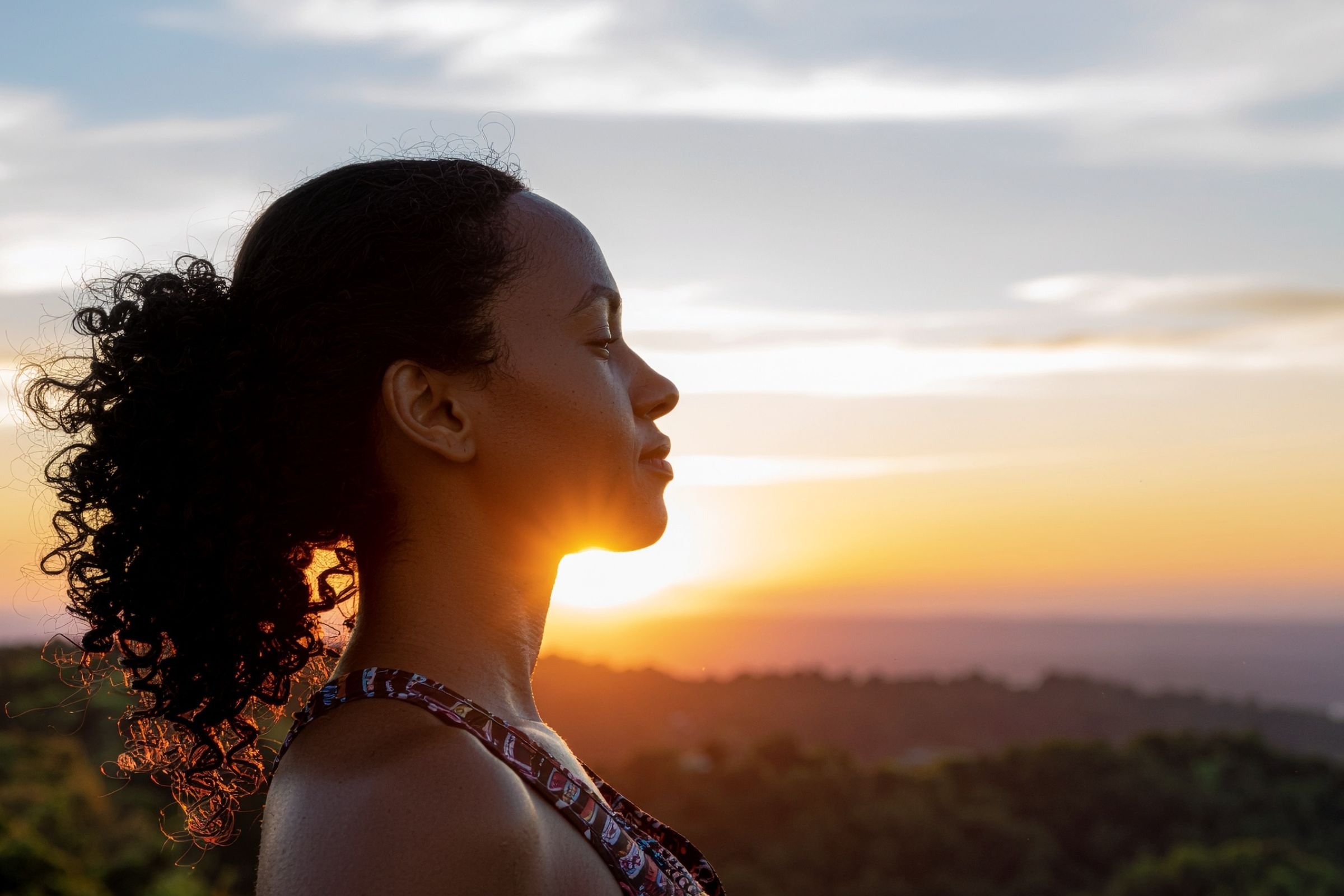 A woman with curly hair closes her eyes in a serene pose, silhouetted against a vibrant sunset. The scene conveys peace and tranquility.