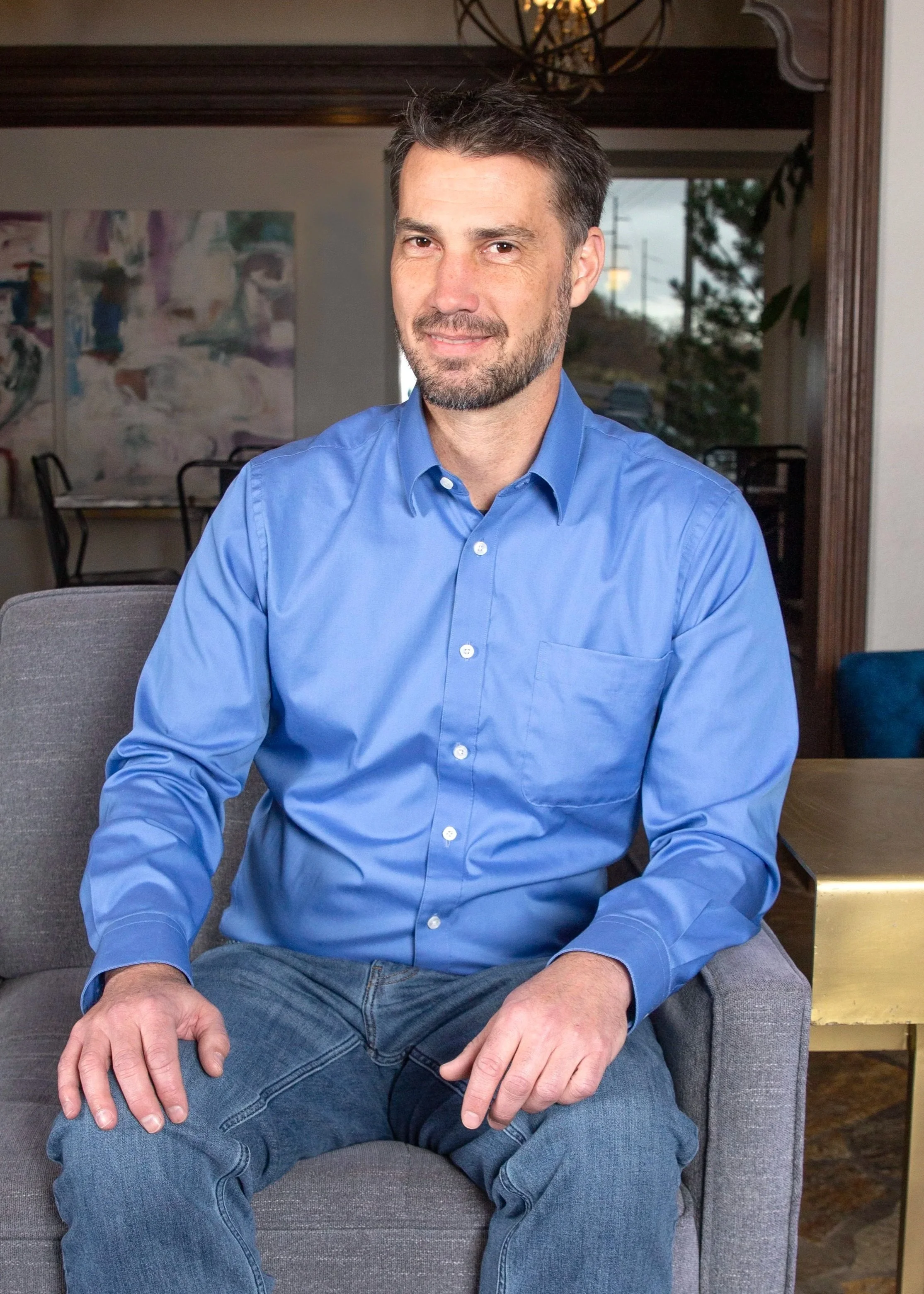 Man with short dark hair and light stubble, wearing a blue button-down shirt and jeans, seated on a gray armchair indoors and smiling slightly at the camera.