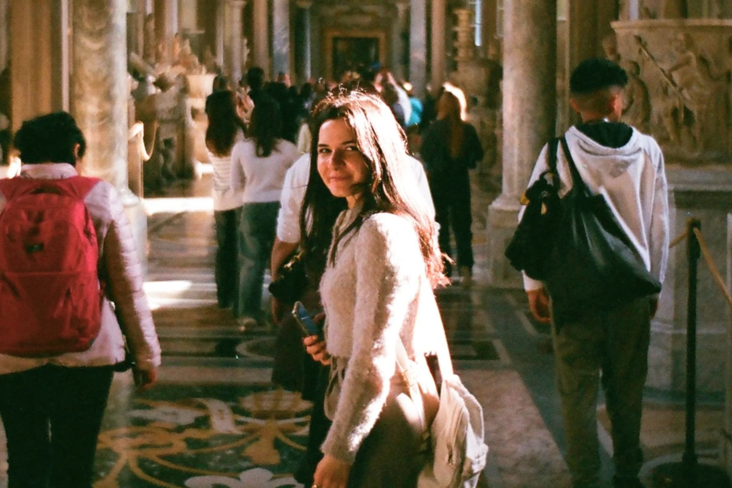 A smiling student with long brown hair looks back over her shoulder while walking through a grand, sunlit museum gallery lined with classical stone columns and distant visitors.
