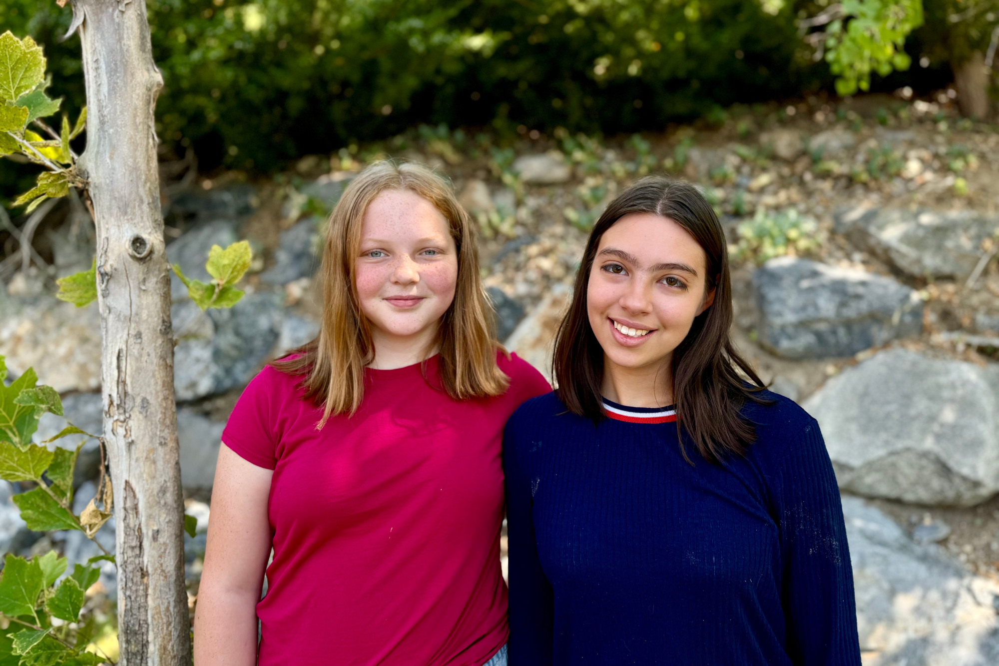 Two students smiling outdoors in front of rocks and greenery.