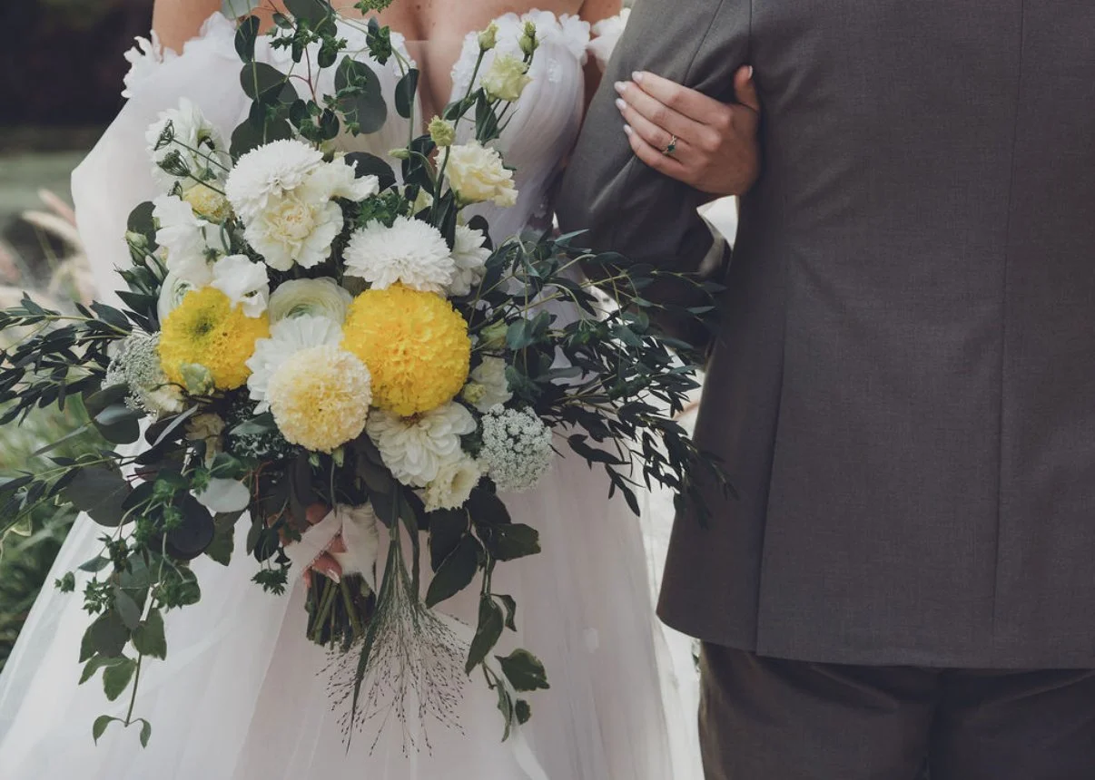 A bride holding a large bouquet of white and yellow flowers, with a groom standing beside her, during a wedding ceremony.
