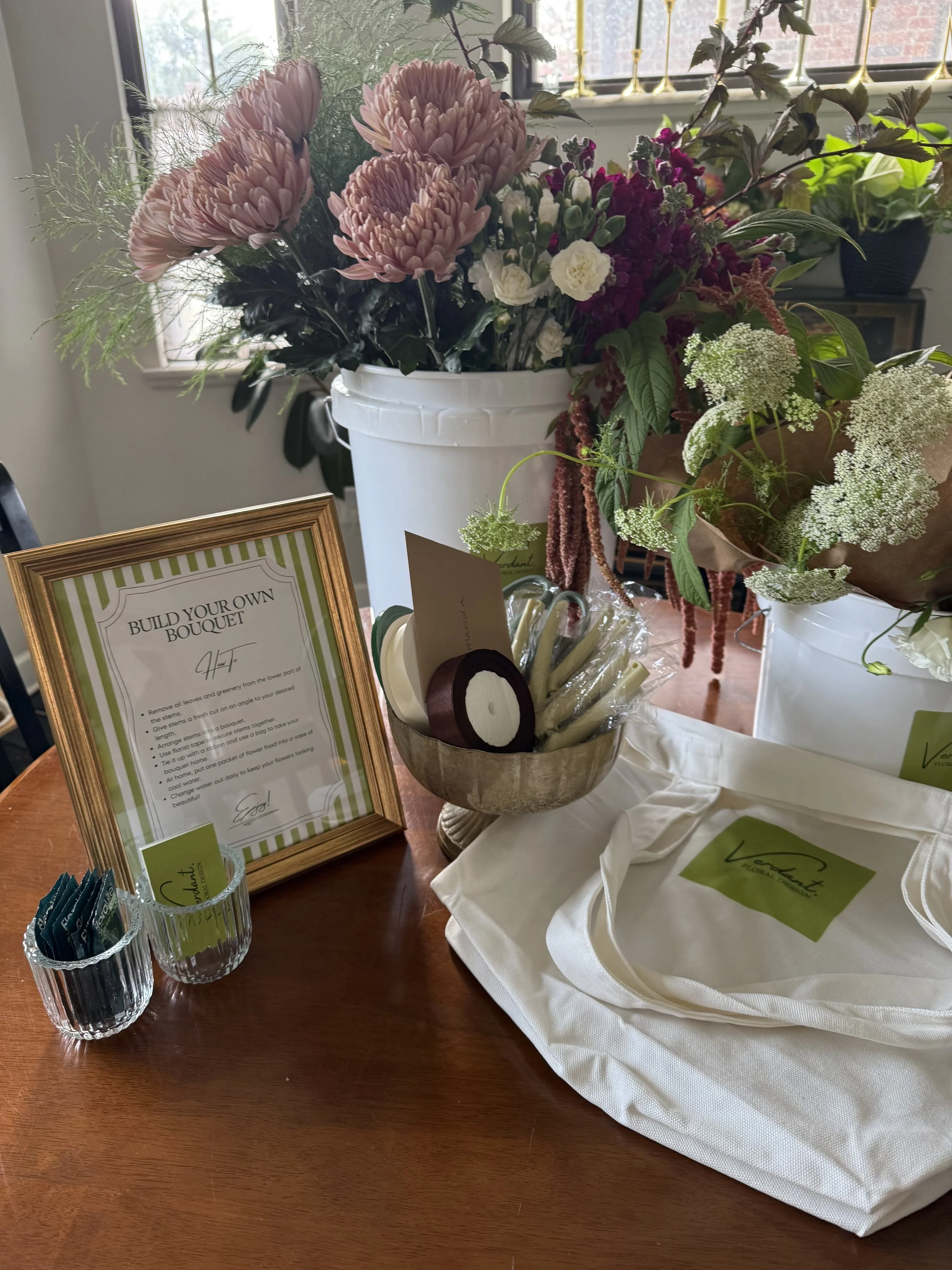 Floral arrangement with pink, white, and green flowers in a white bucket on a wooden table, with a frame displaying instructions, small containers with tea bags, and a cloth bag with a green label.