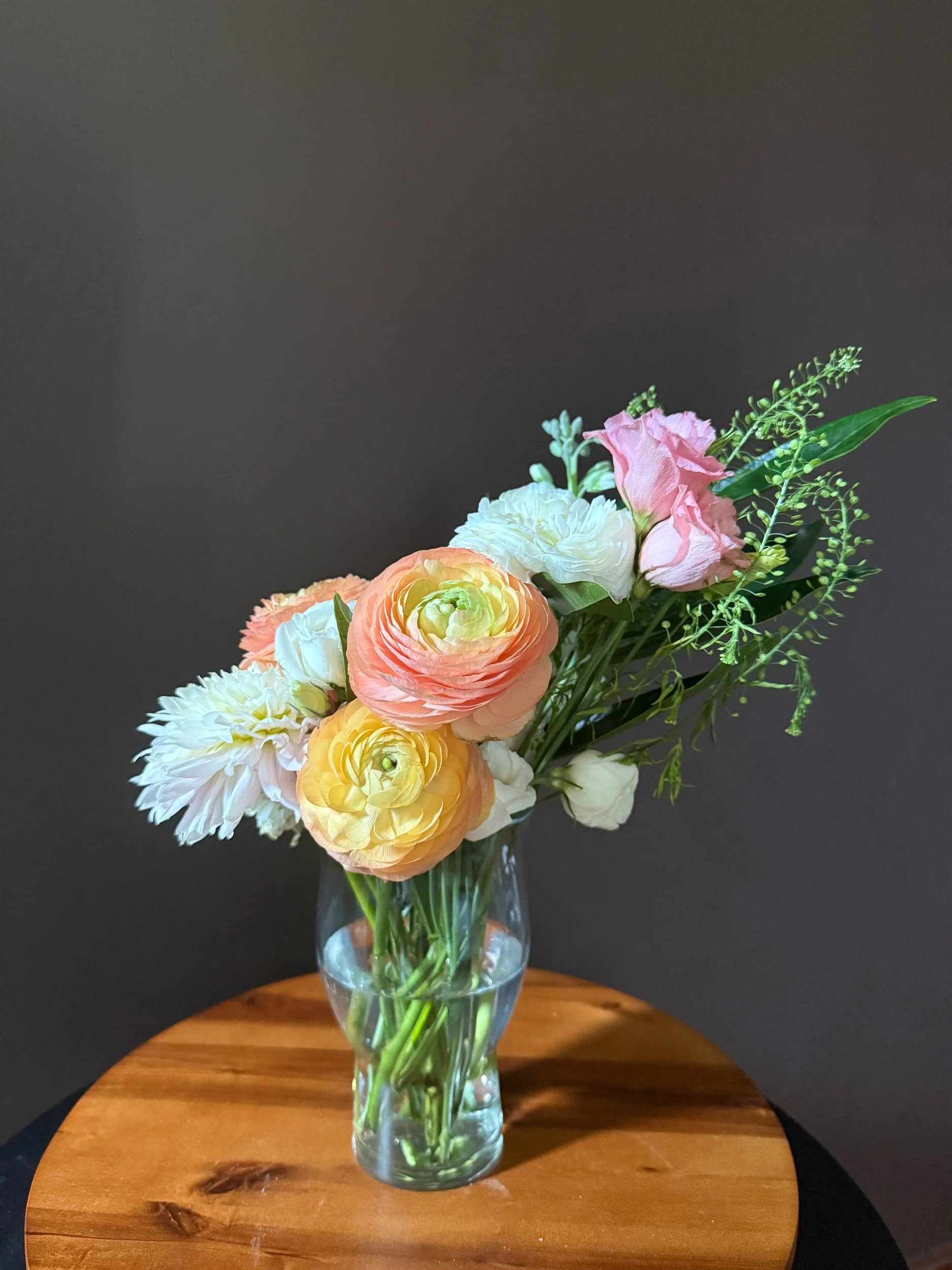 A glass vase with a bouquet of mixed flowers, including pink, peach, white, and yellow blossoms, is placed on a wooden surface against a dark background.