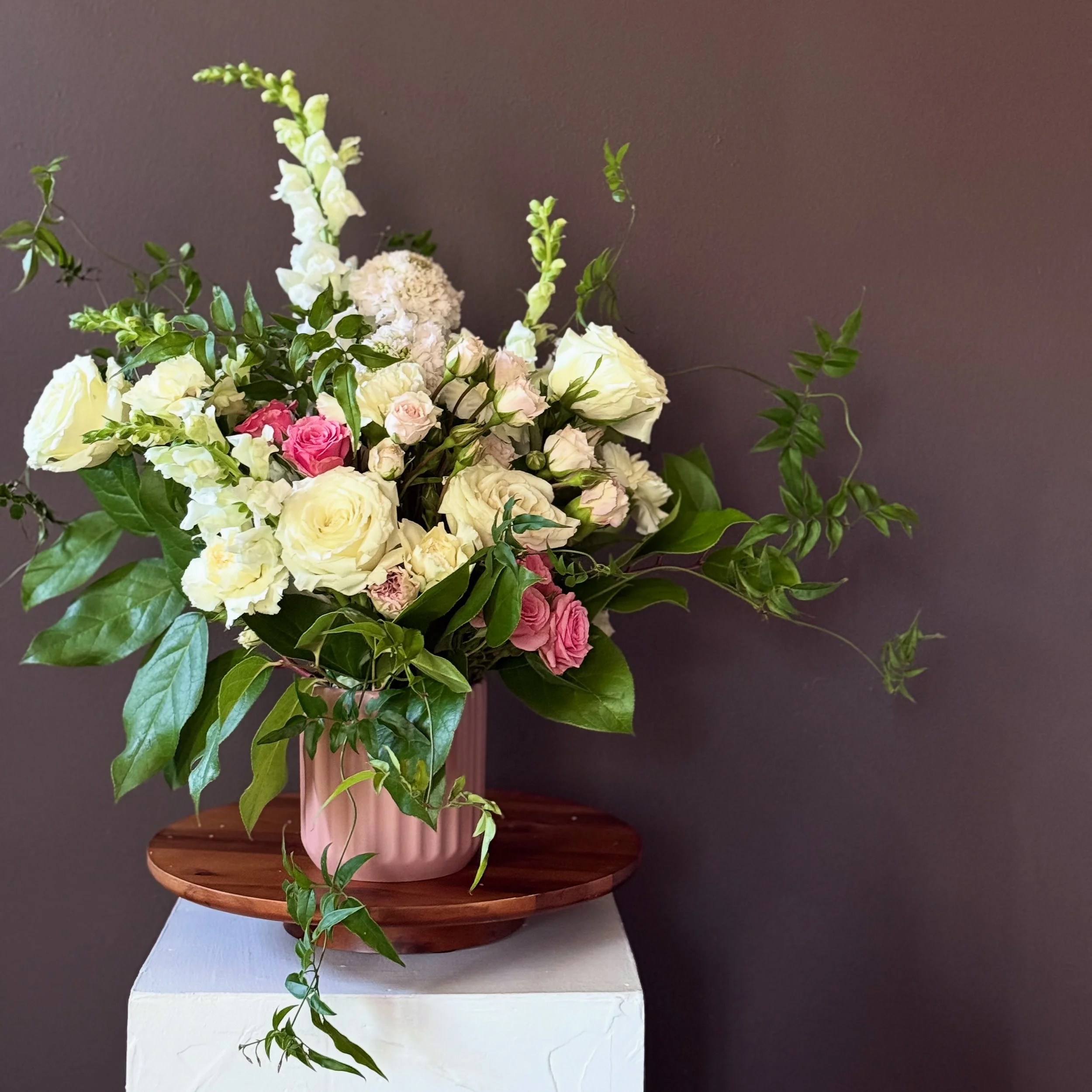 A bouquet of white and pink roses with green foliage in a pink striped vase on a wooden tray against a purple wall.
