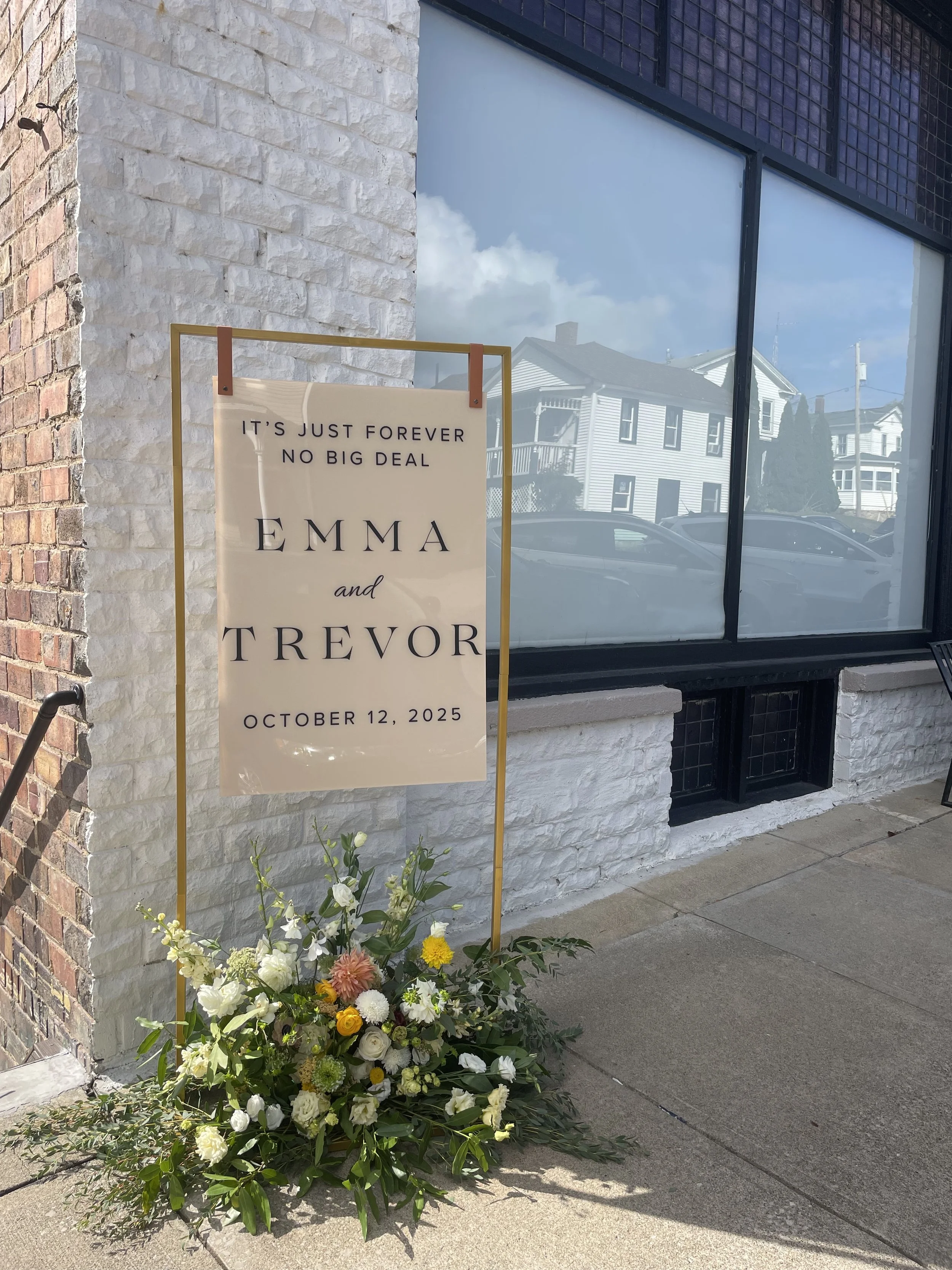 Wedding sign with flowers outside a building. The sign reads, 'It's just forever, no big deal, Emma and Trevor, October 12, 2025.'